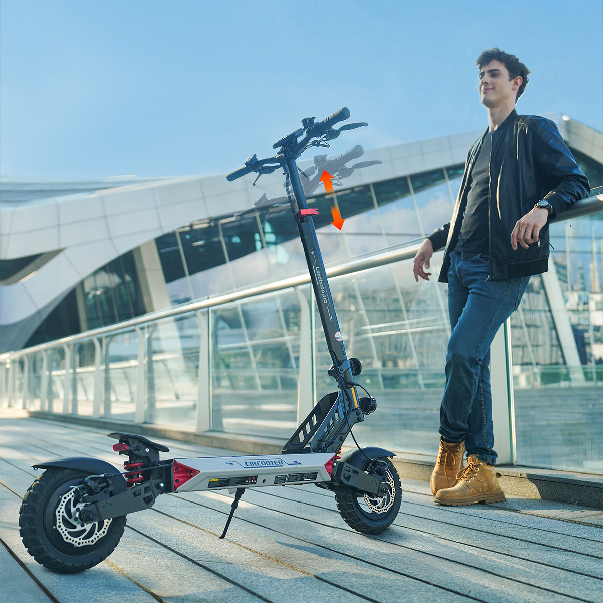 Mężczyzna opiera się o barierkę obok hulajnogi elektrycznej. Hulajnoga jest czarna i szara. Tło to nowoczesny budynek.