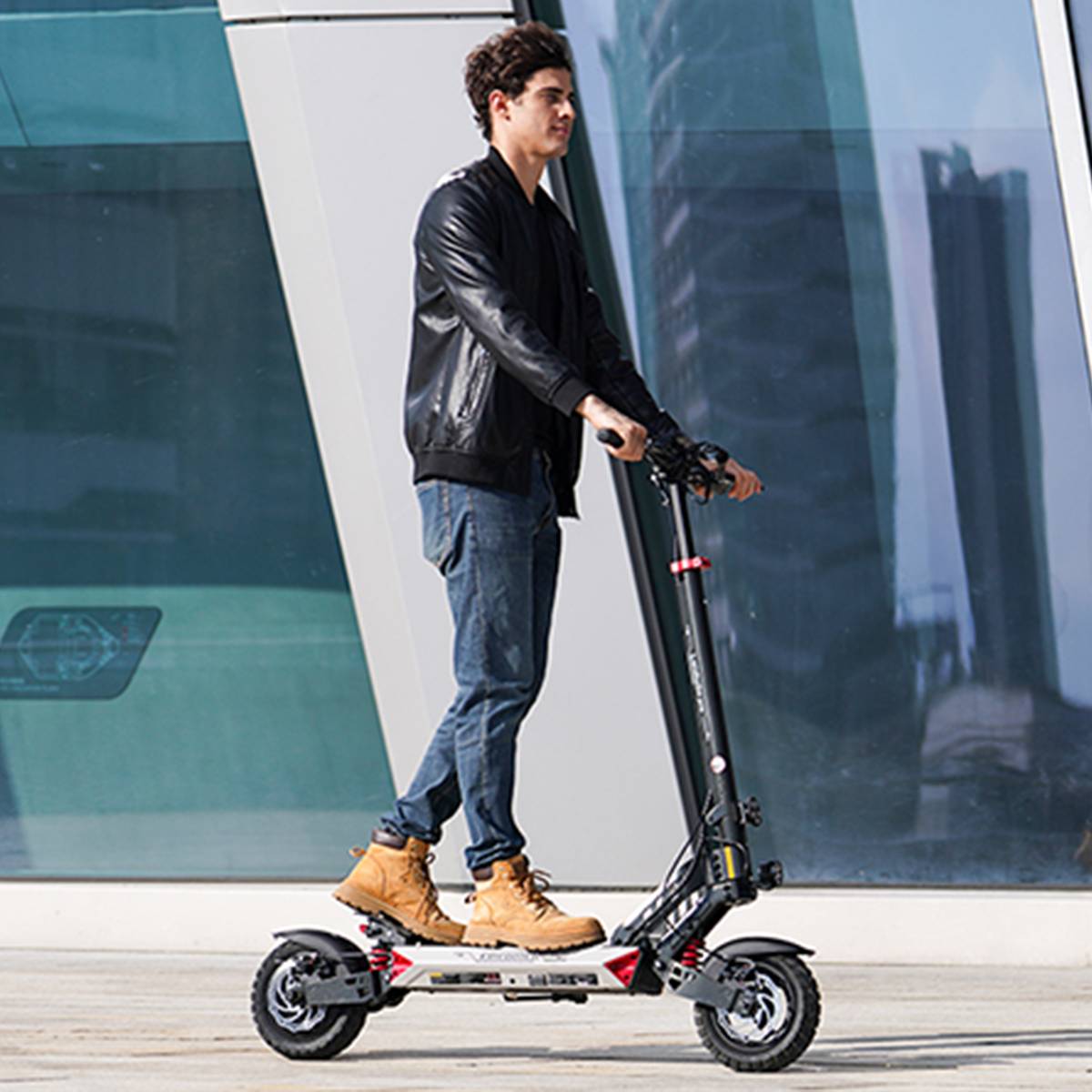 Mężczyzna w czarnej skórzanej kurtce i dżinsach jedzie na hulajnodze elektrycznej. Ma brązowe buty. Tło to nowoczesny budynek.