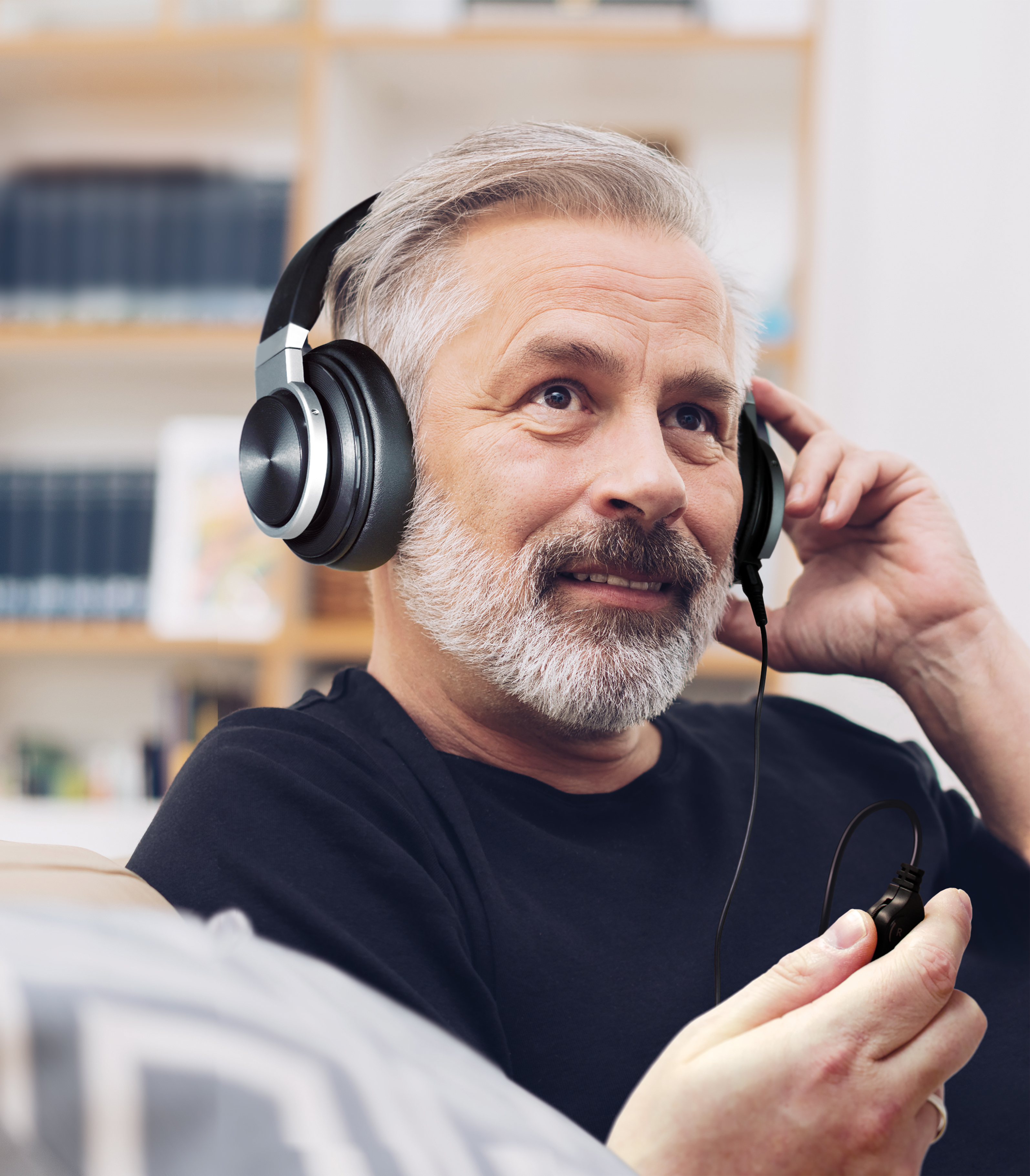 Ein Mann mit Kopfhörern lächelt und hält das Kabel. Er trägt Schwarz und sitzt. Bücherregal im Hintergrund.