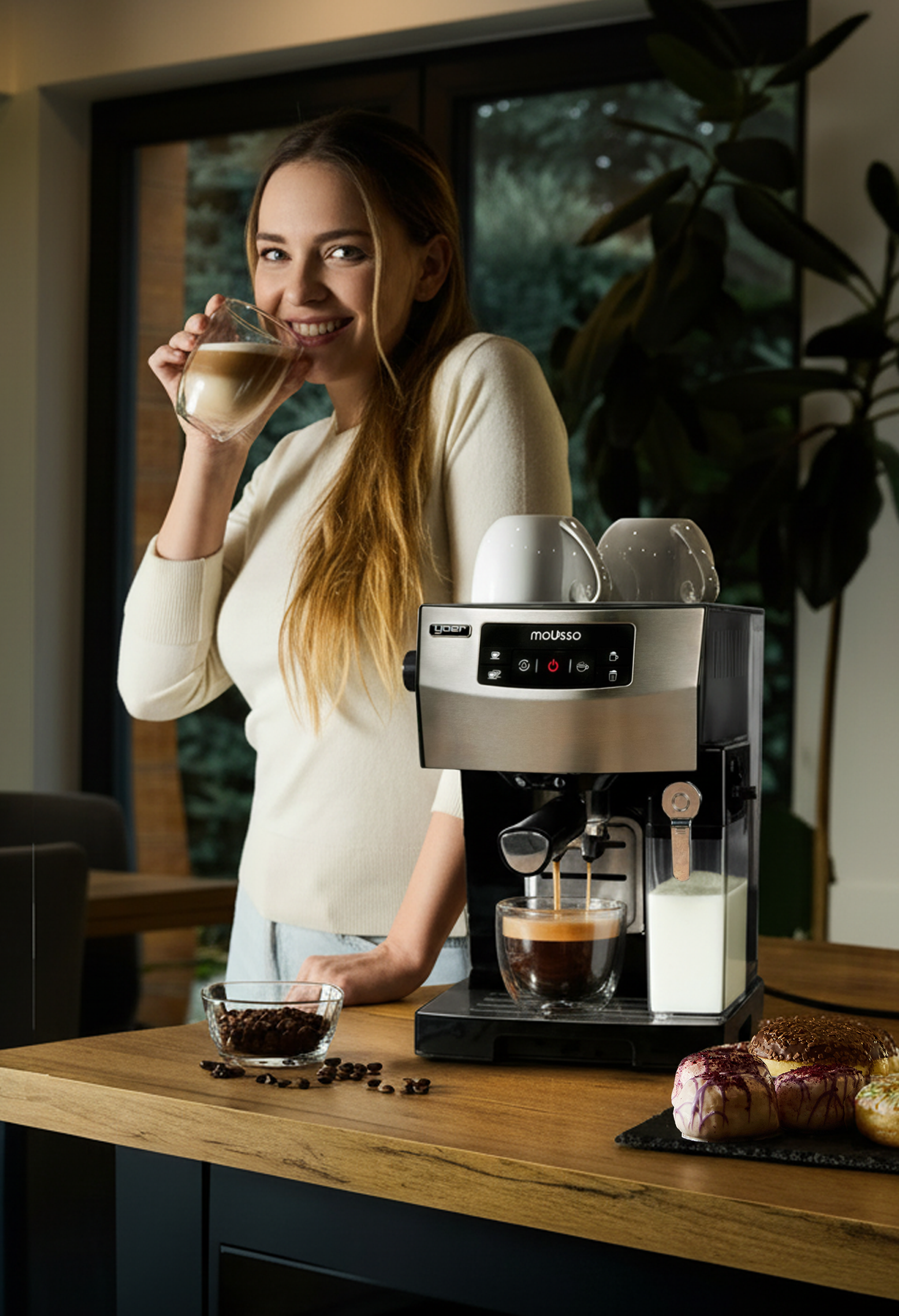 Kobieta pije kawę obok ekspresu do kawy. Na stole jest miska z ziarnami i pączki.