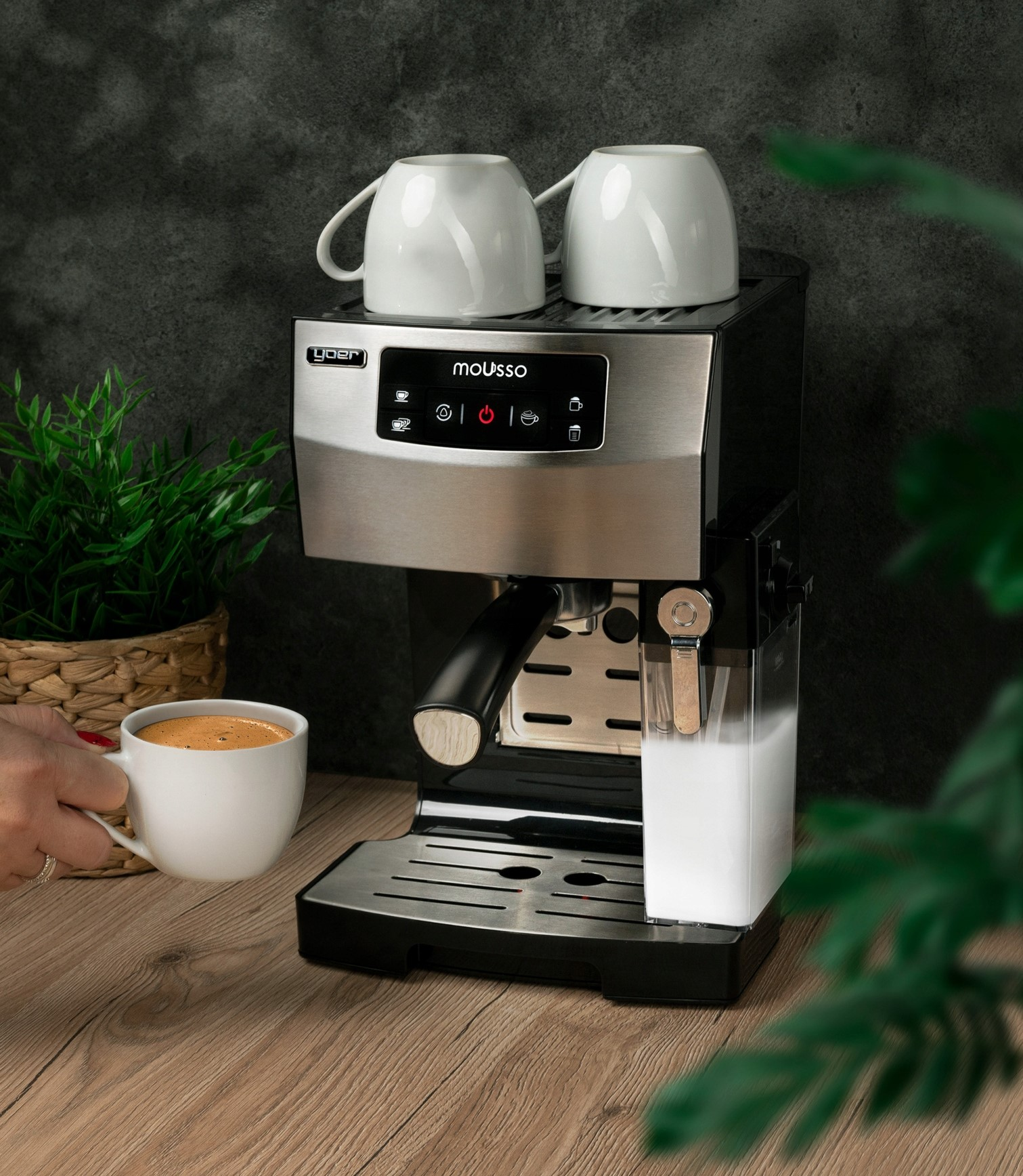 Ekspres do kawy z dwoma filiżankami na górze, filiżanką kawy i pojemnikiem na mleko. Ręka trzyma filiżankę kawy.