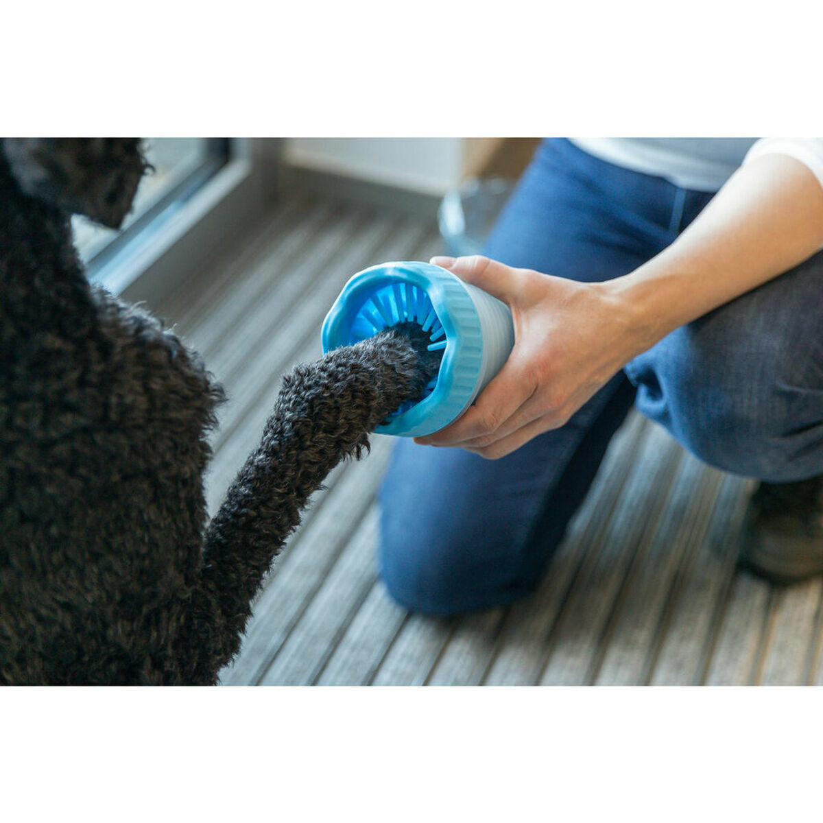 Una persona tiene in mano un pulitore per zampe blu e bianco, pulendo la zampa nera di un cane. Sono visibili il corpo del cane e le gambe di una persona.
