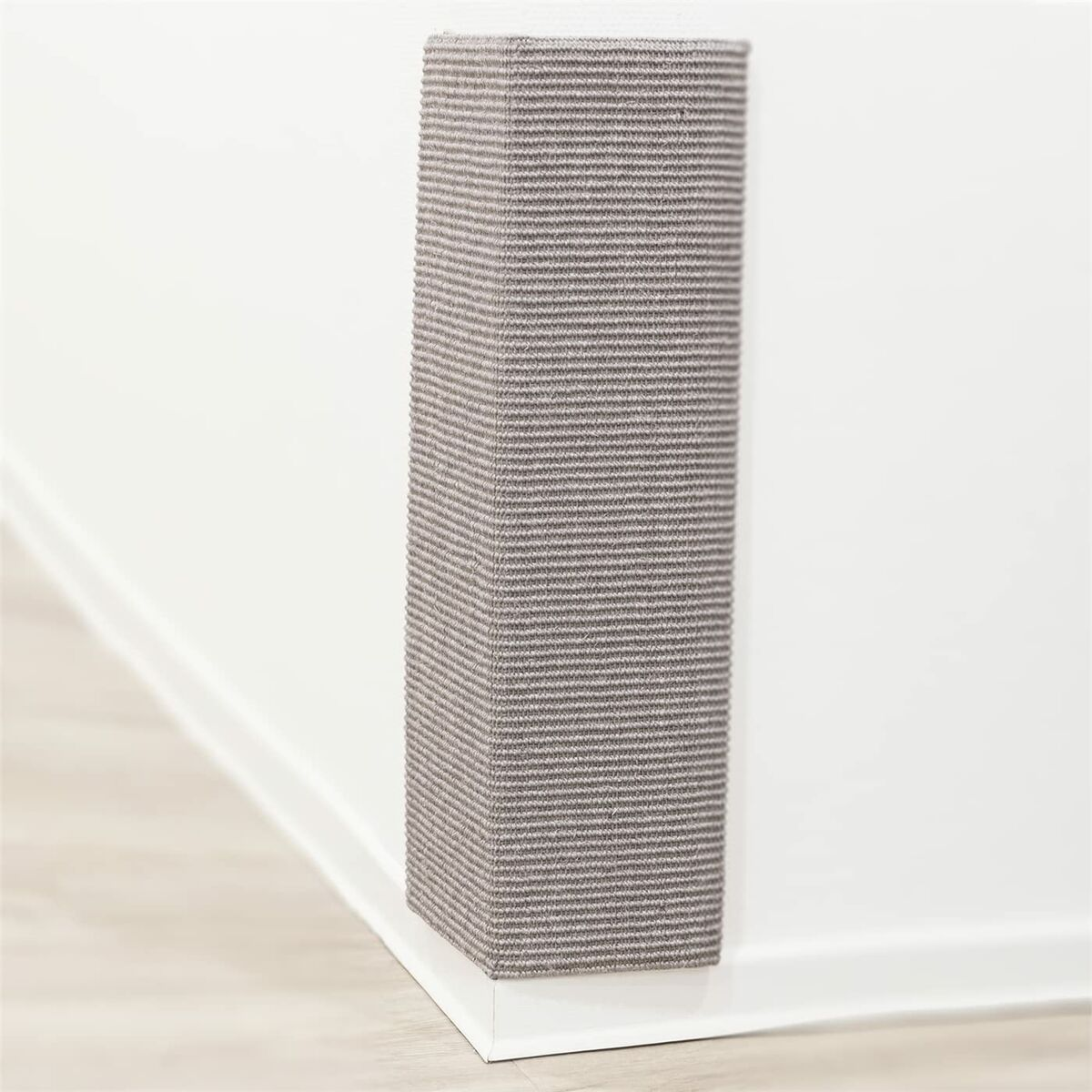 Un rascador para gatos gris y rectangular está contra una pared blanca y un zócalo blanco. El suelo es de madera clara.