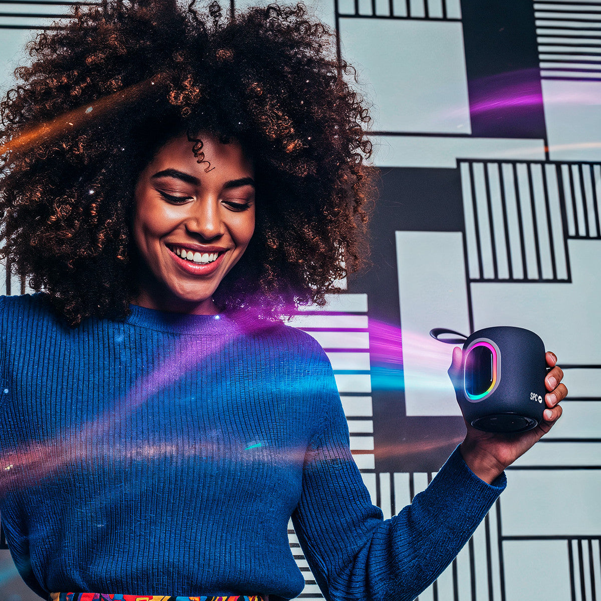 Donna sorridente tiene in mano un altoparlante nero con luci colorate. Indossa un maglione blu su sfondo geometrico bianco e nero.