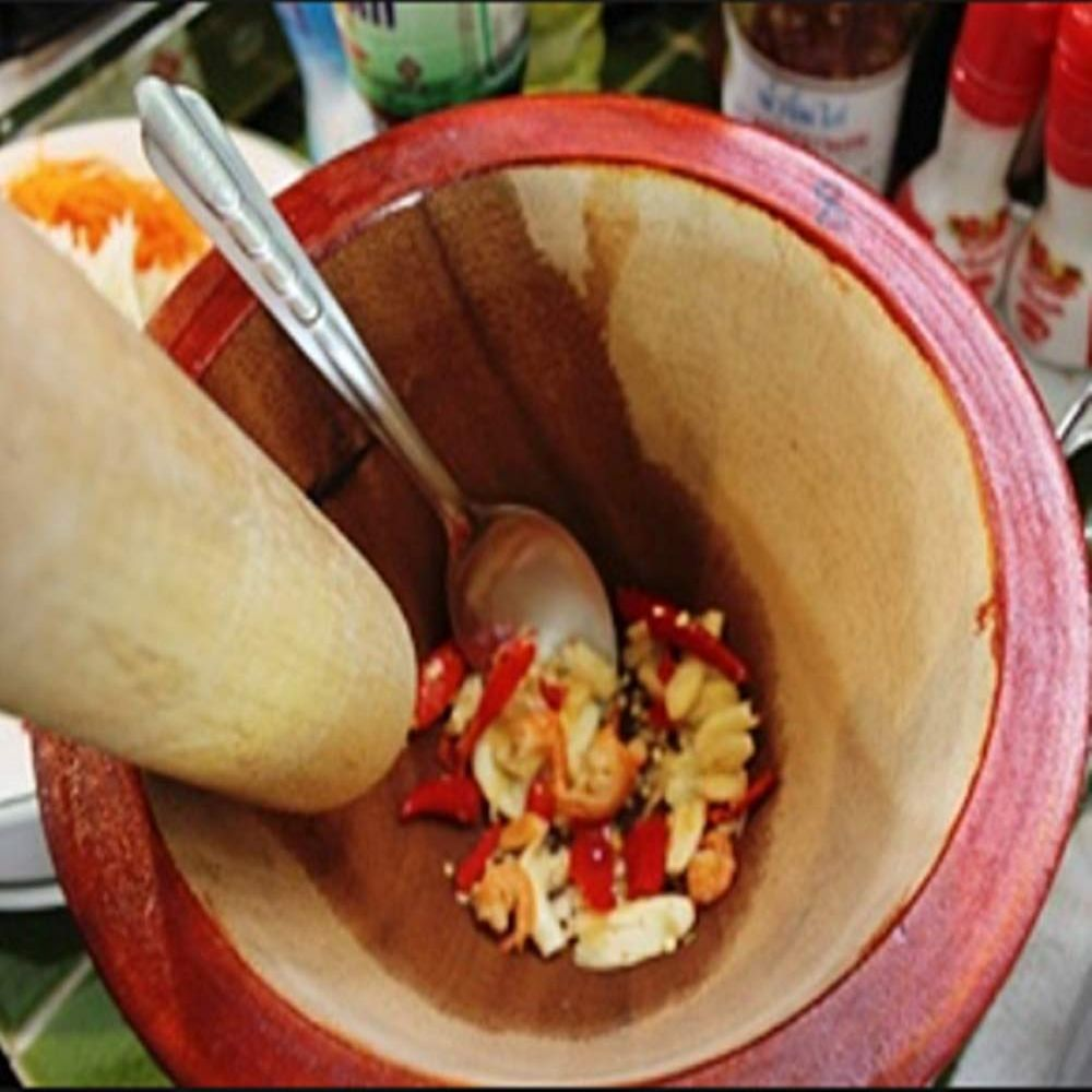 Holzmörser mit Zutaten, Löffel und Stößel. Rote Chilis, Garnelen und Knoblauch innen. Weitere Zutaten und Flaschen im Hintergrund.