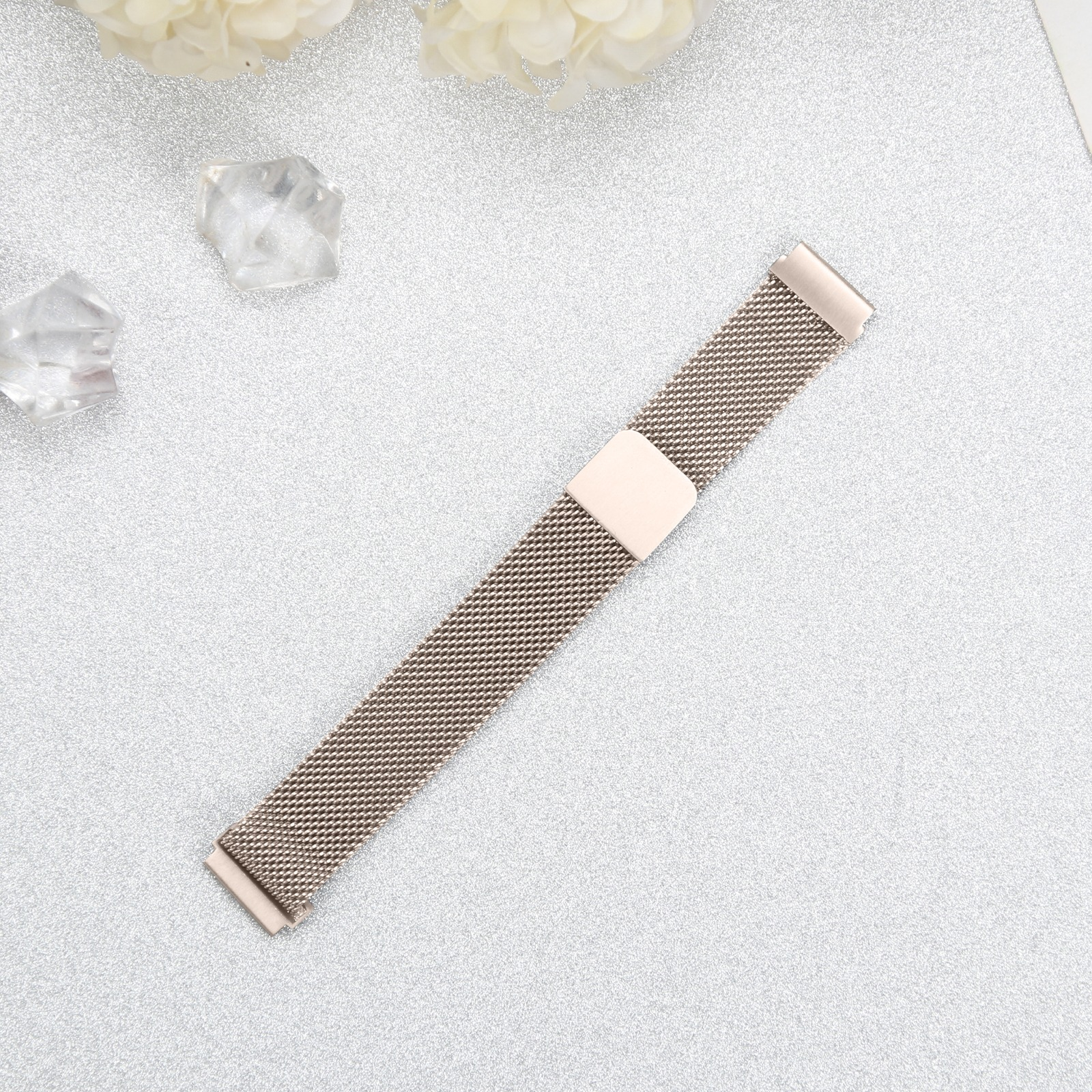 Ein roségoldenes Uhrenarmband auf einer glitzernden silbernen Oberfläche, mit klaren kristallartigen Objekten und weißen Blumen.