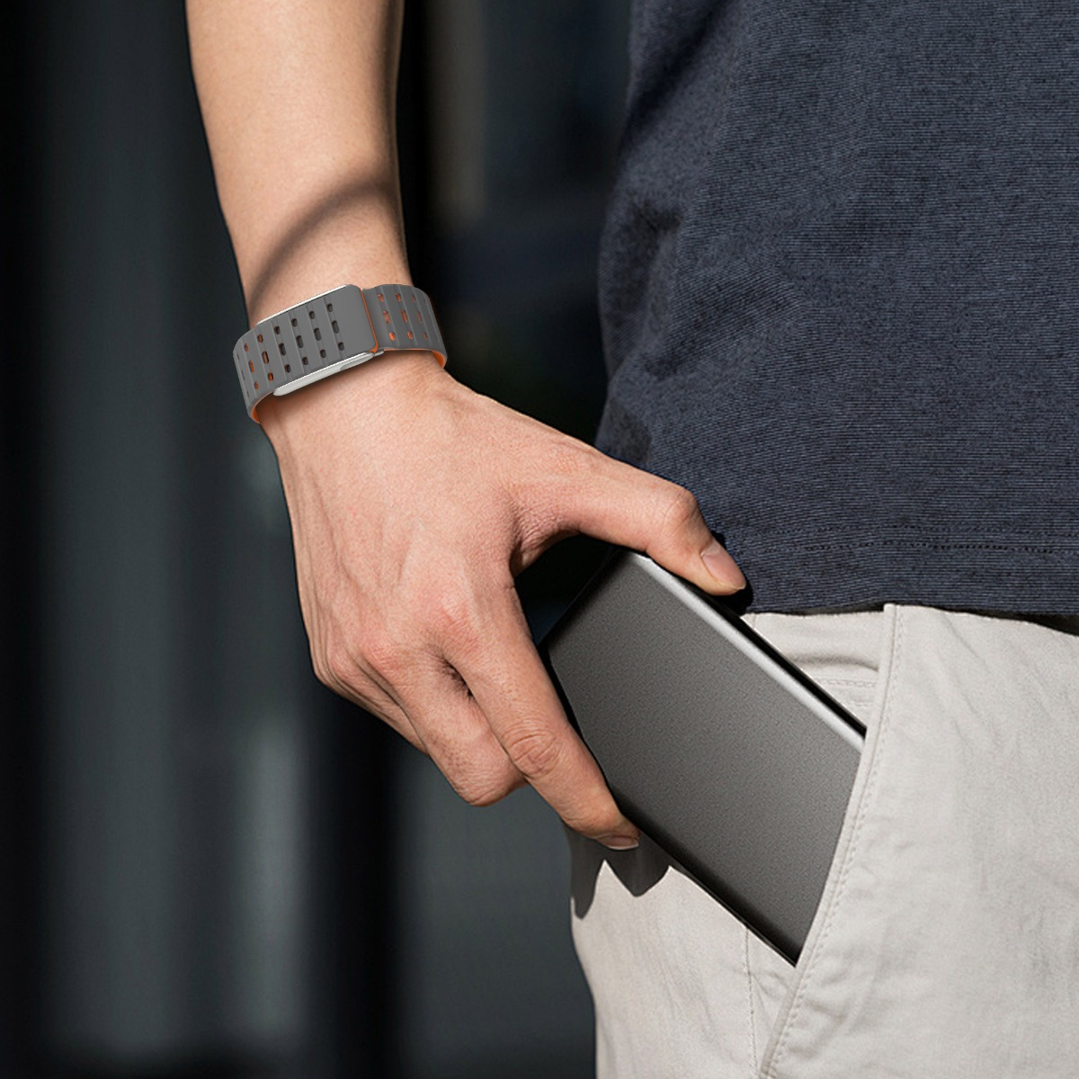 Eine Person mit einer grauen Smartwatch und einem grauen Hemd, die ein graues Telefon in die Tasche steckt. Der Hintergrund ist verschwommen.