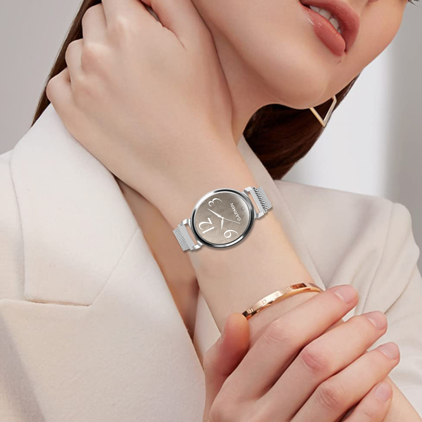 Das Handgelenk einer Frau mit einer Garmin-Uhr. Die Uhr hat ein silbernes Armband und ein beiges Zifferblatt. Die Frau trägt ein goldenes Armband.