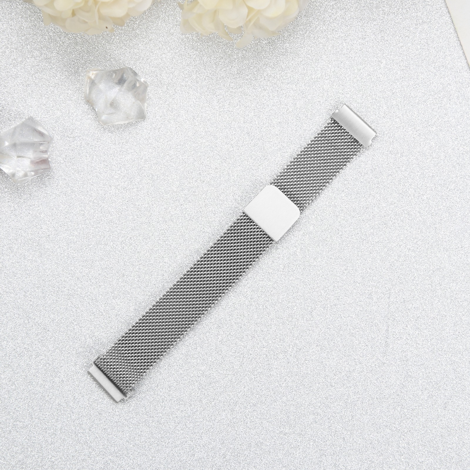 Ein silbernes Mesh-Uhrenarmband mit weißem Verschluss liegt auf glitzernder Silberoberfläche, mit Blumen und Kristallen.