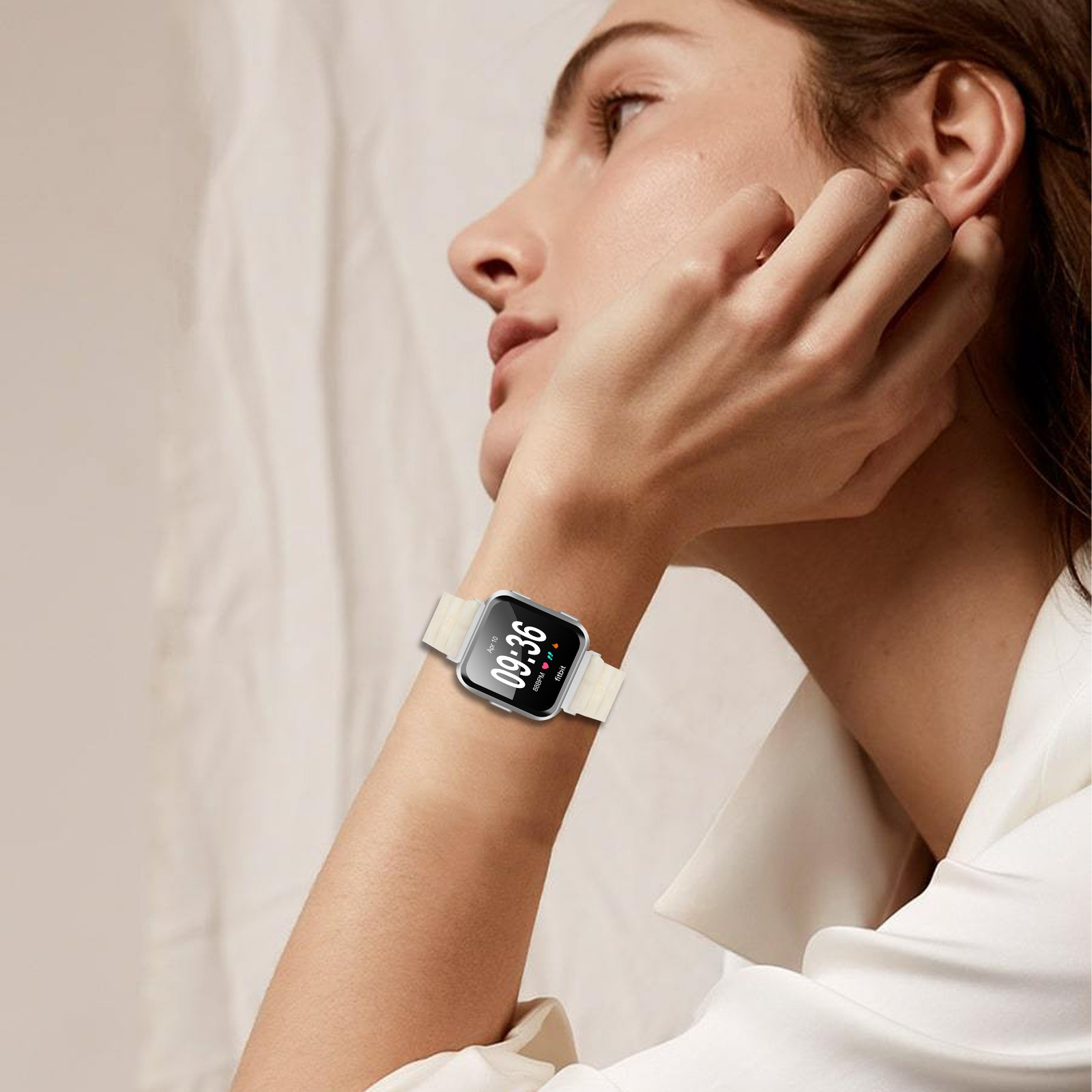 Eine Frau trägt eine Smartwatch. Die Uhr zeigt die Zeit an. Sie berührt ihr Ohr. Cremefarbenes Uhrenarmband.