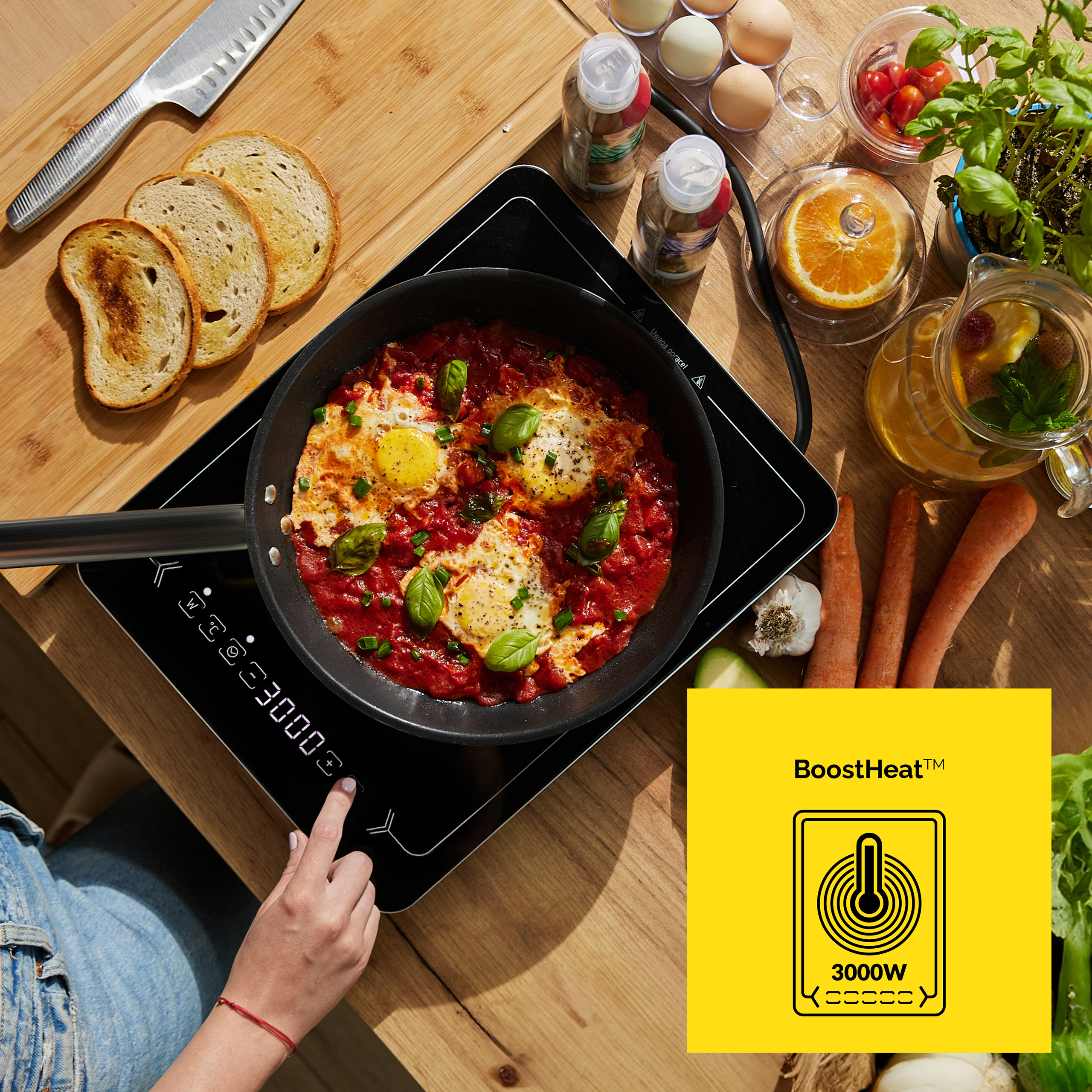 Patelnia z jajkami w sosie pomidorowym na kuchence. Widoczne są również chleb, warzywa i dłoń dotykająca elementów sterujących.