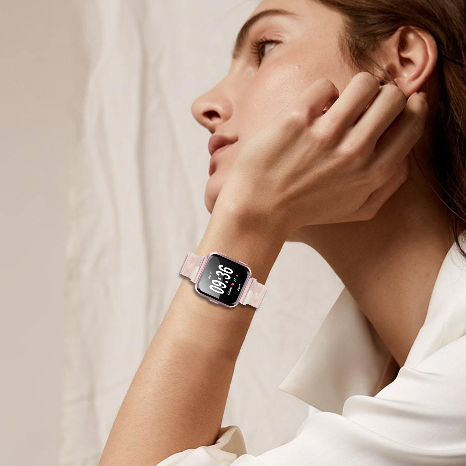 Una donna indossa uno smartwatch che mostra 09:36. L'orologio ha un cinturino rosa. Si tocca l'orecchio.