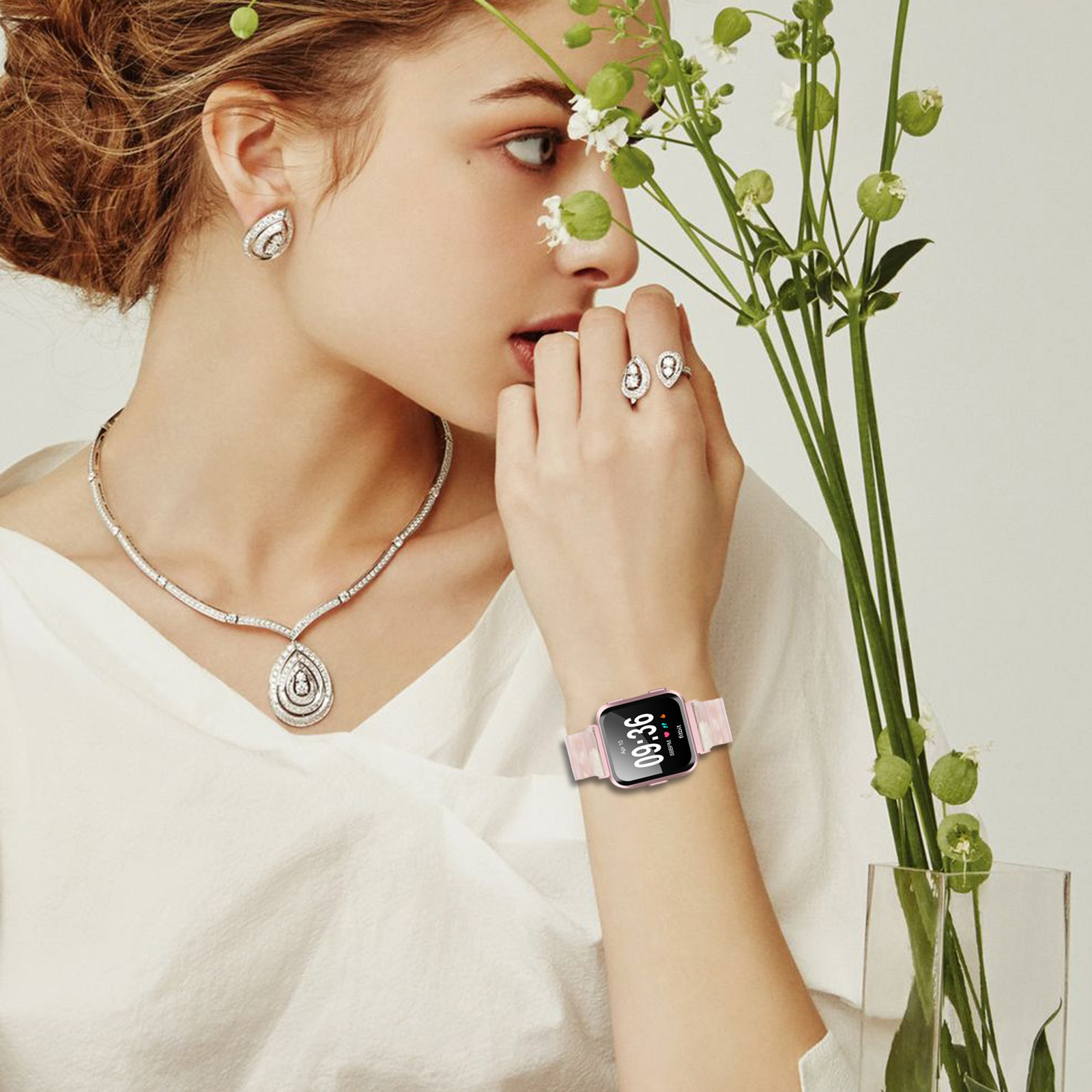 Donna con gioielli e smartwatch, tiene dei fiori. Ha orecchini, collana e anelli. L'orologio segna le 09:36.