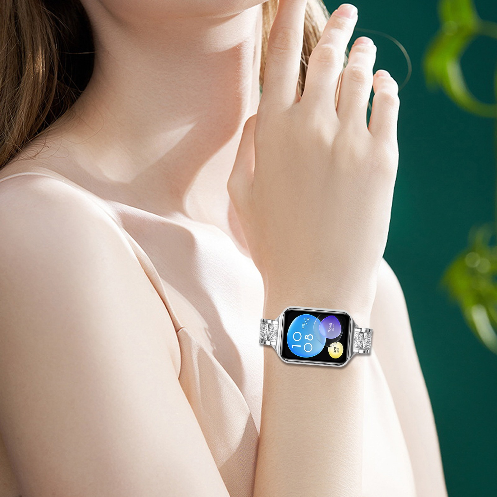 Il braccio di una donna con uno smartwatch. L'orologio ha una cornice argentata e un cinturino gioiello. Lo sfondo è verde.