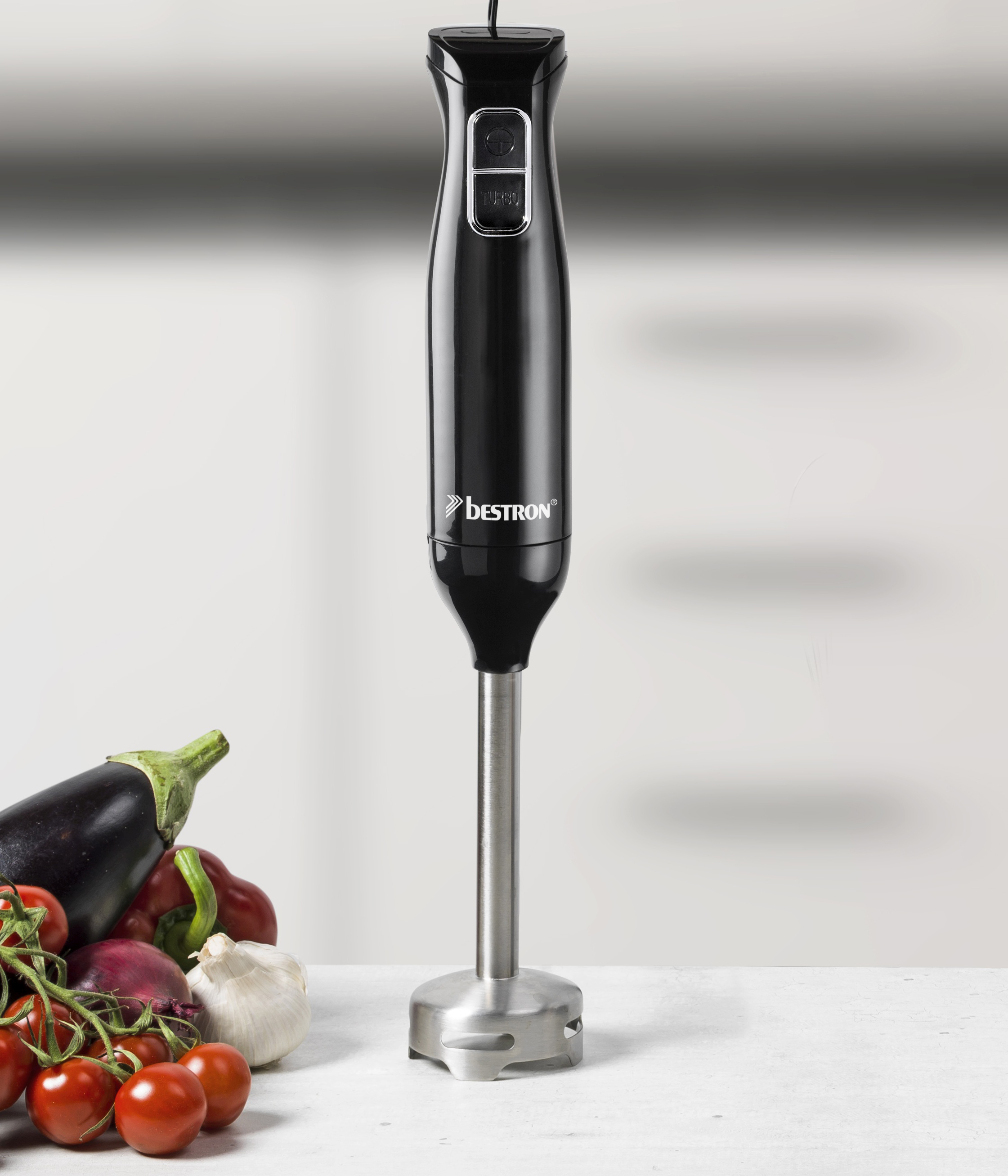 Un frullatore a immersione nero sopra le verdure. Pomodori, melanzane, cipolla e aglio su una superficie bianca.