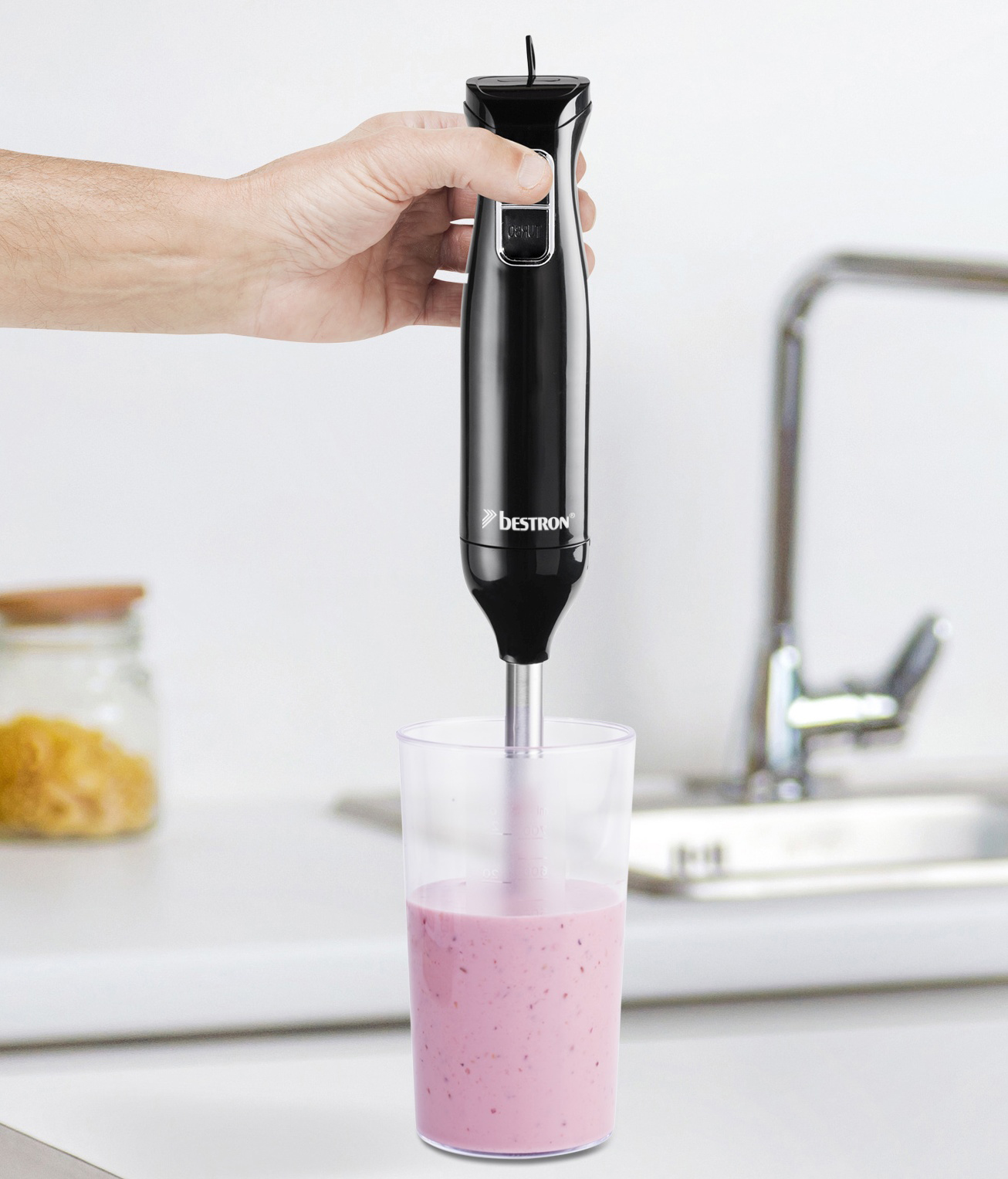 Una mano tiene un frullatore a immersione nero sopra un bicchiere di frullato rosa. Sfondo cucina con lavandino e barattolo.