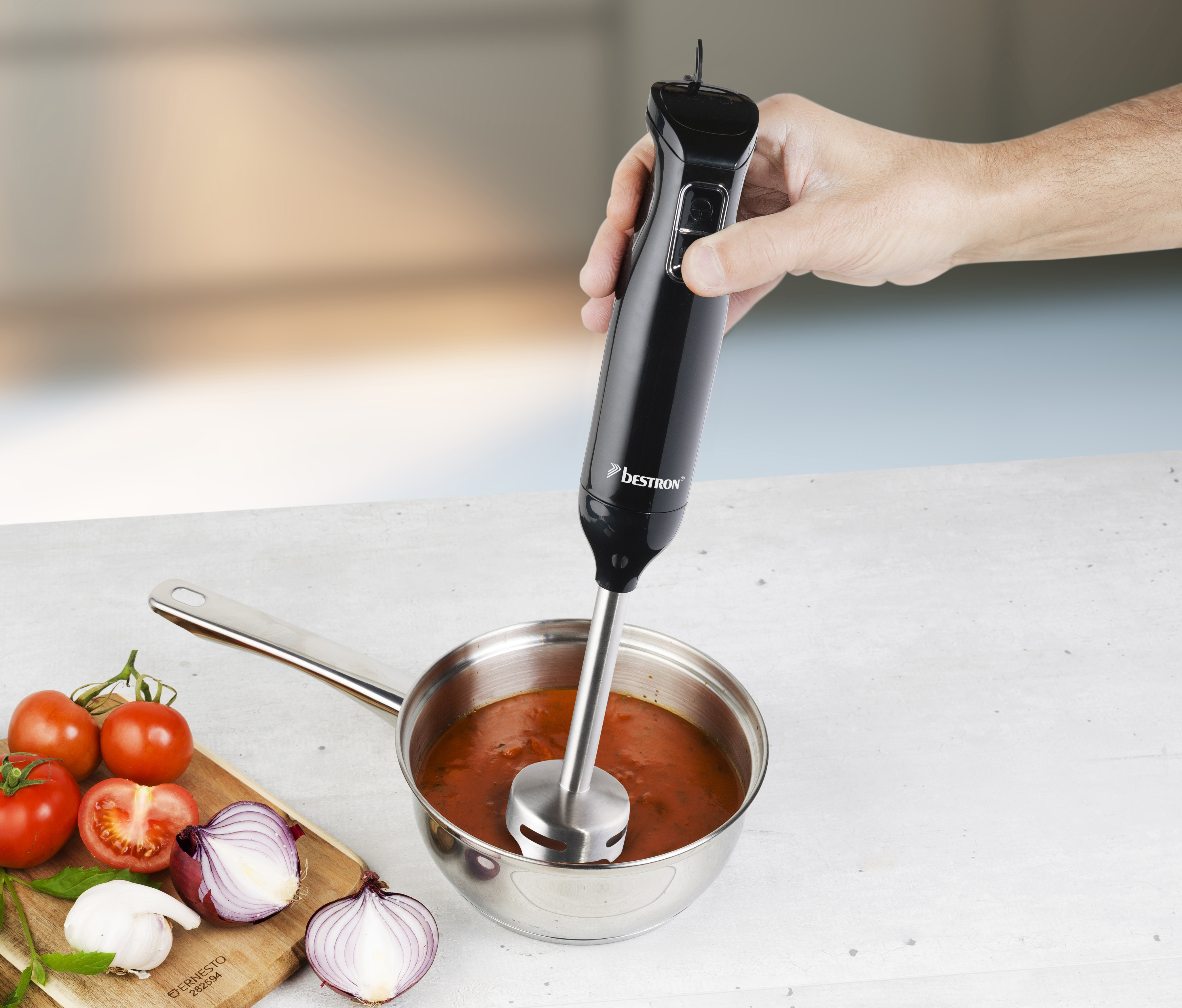 Una mano che tiene un frullatore a immersione nero frulla la zuppa in una pentola. Pomodori, cipolle e aglio sono di lato.