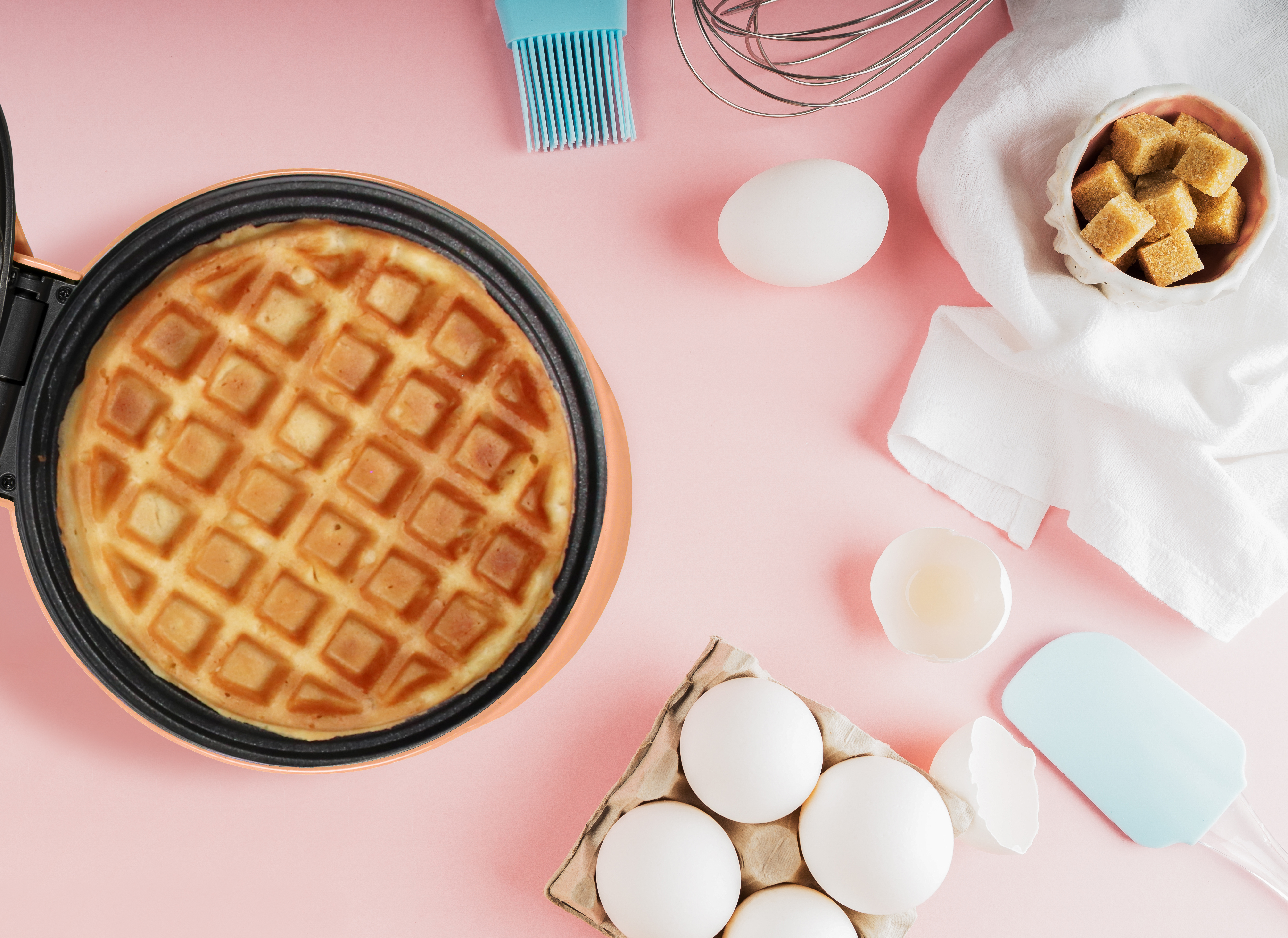 Waffle in a waffle iron, eggs, sugar cubes, whisk, and spatula on a pink surface.