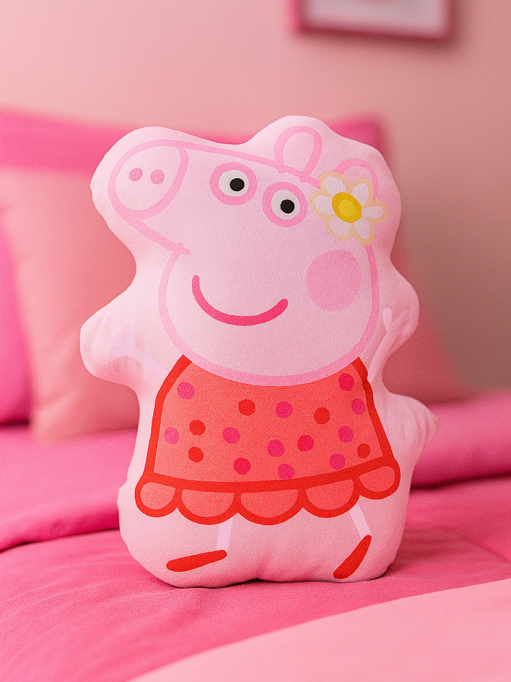 Rosa Peppa Wutz Kissen auf einem rosa Bett. Peppa trägt ein rotes Kleid mit pinken Punkten. Eine Blume schmückt ihren Kopf.