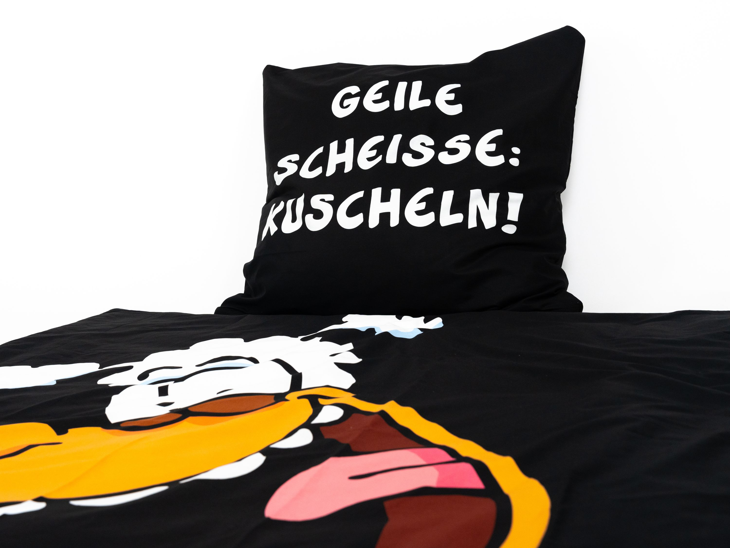 Schwarzes Kissen mit weißem Text 'GEILE SCHEISSE: KUSCHELN!' und ein schwarzes Laken mit einem Cartoon.