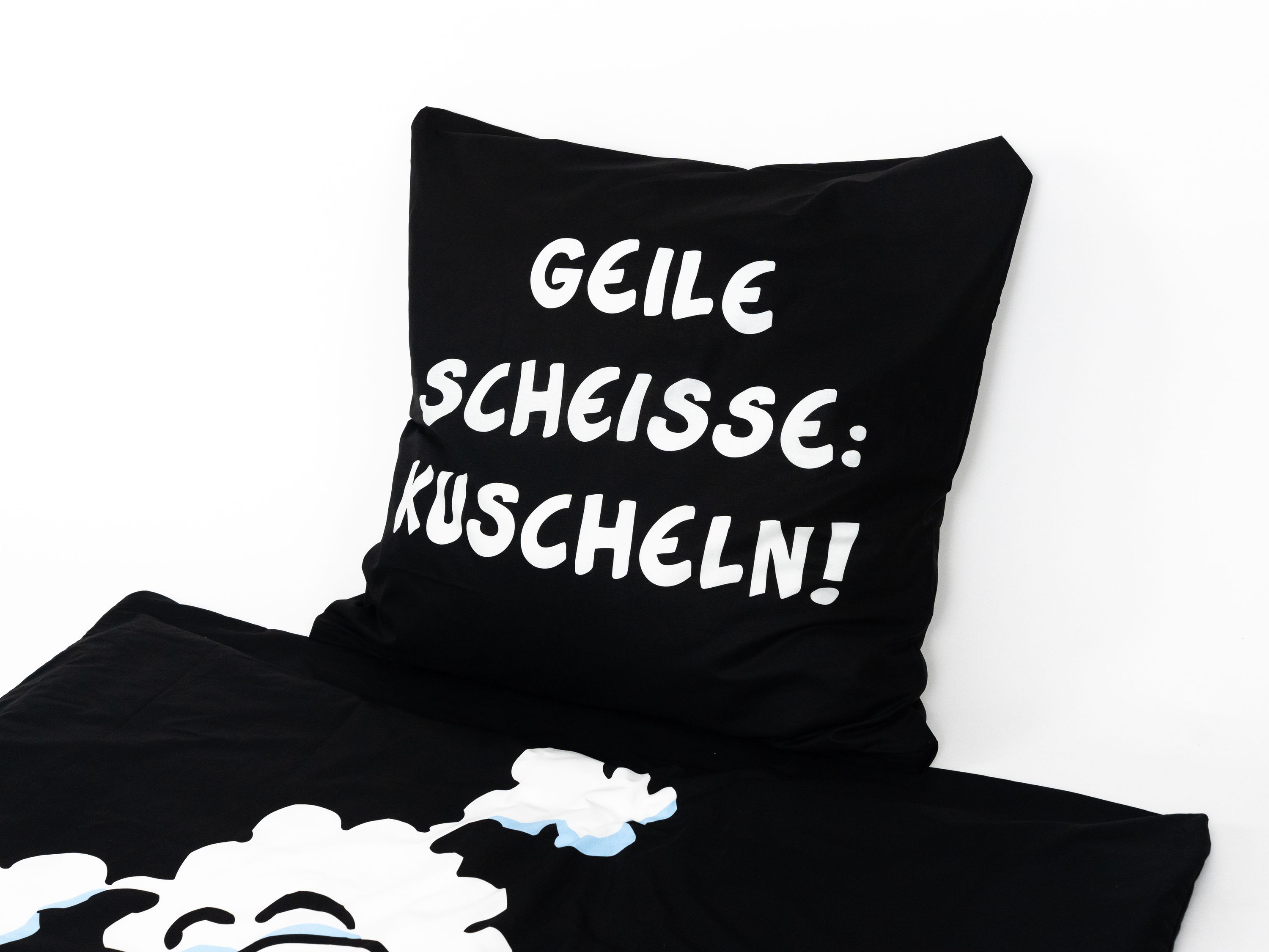 Schwarzes Kissen mit weißem Text: GEILE SCHEISSE: KUSCHELN! Unten: schwarzes Laken mit weißen und hellblauen Formen.
