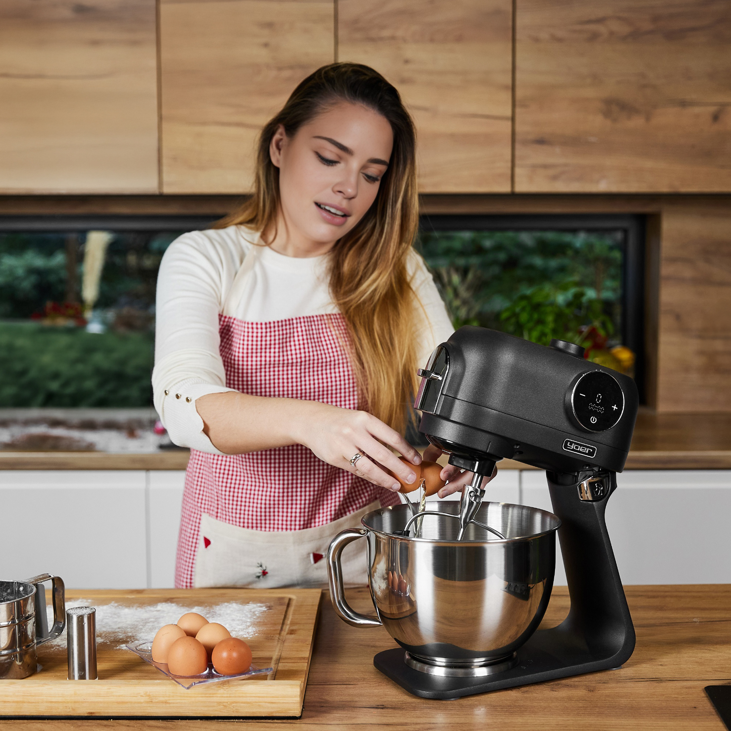 Kobieta wbija jajka do miski miksera. Kuchnia z drewnianymi szafkami i deską do krojenia z jajkami i mąką.