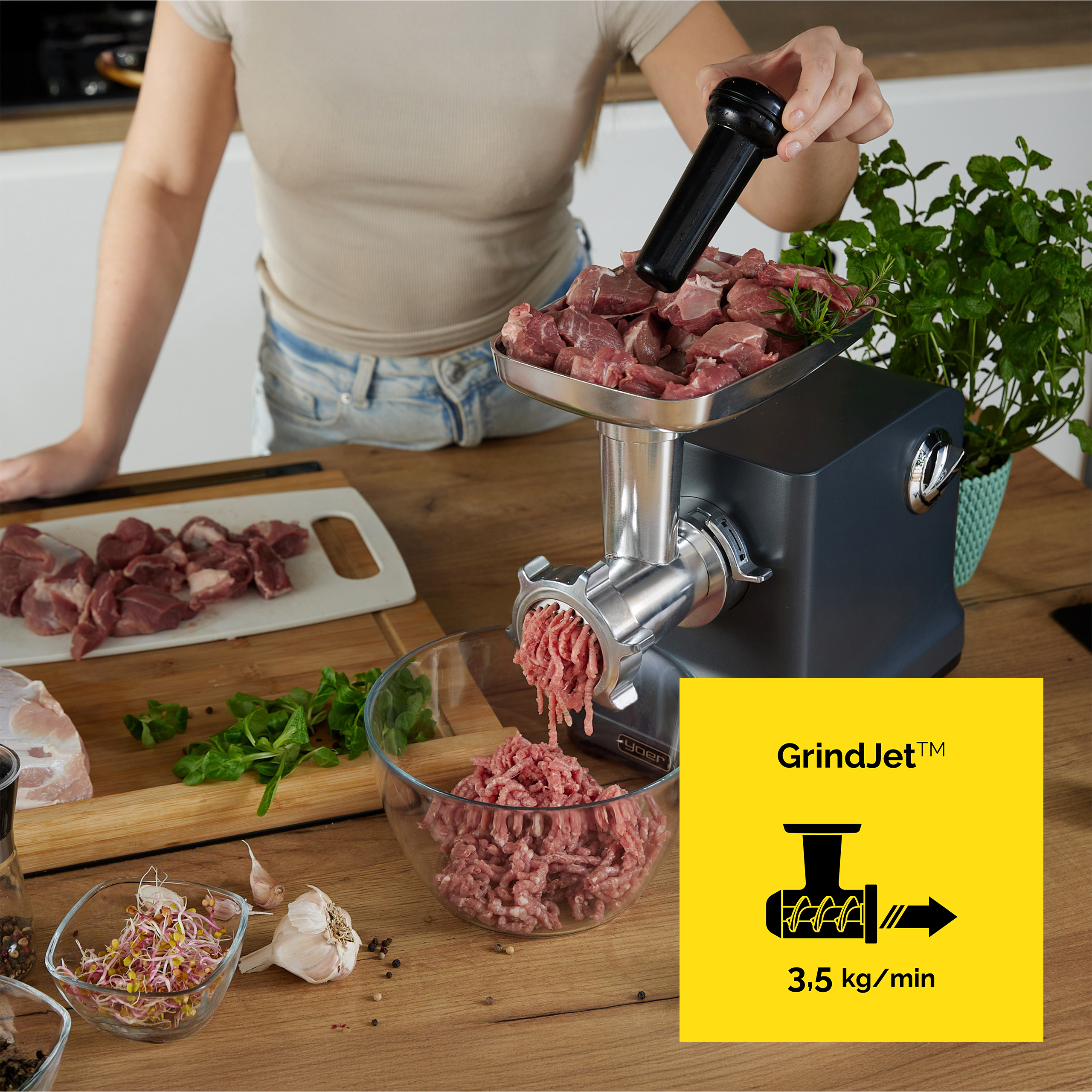 Osoba używa maszynki do mielenia mięsa z surowym mięsem. Mielone mięso wpada do miski. Zioła i składniki na stole.