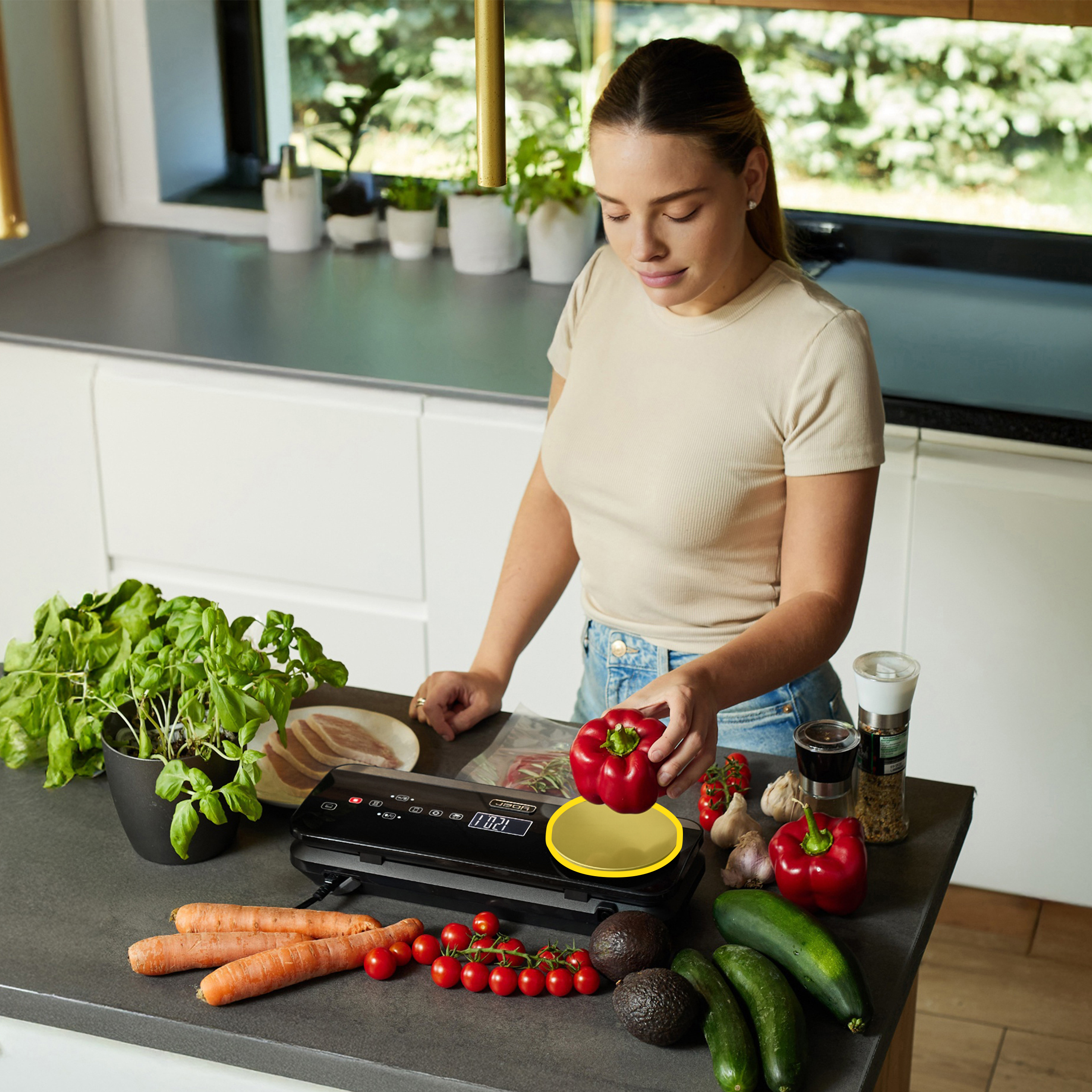 Kobieta trzyma czerwoną paprykę w pobliżu warzyw na blacie kuchennym. Obecne są również zioła i zgrzewarka próżniowa.