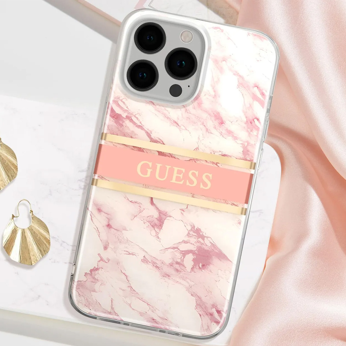 Eine Handyhülle mit rosa Marmormuster und Guess-Logo. Die Hülle liegt auf einer Marmoroberfläche mit Ohrringen und rosa Stoff.
