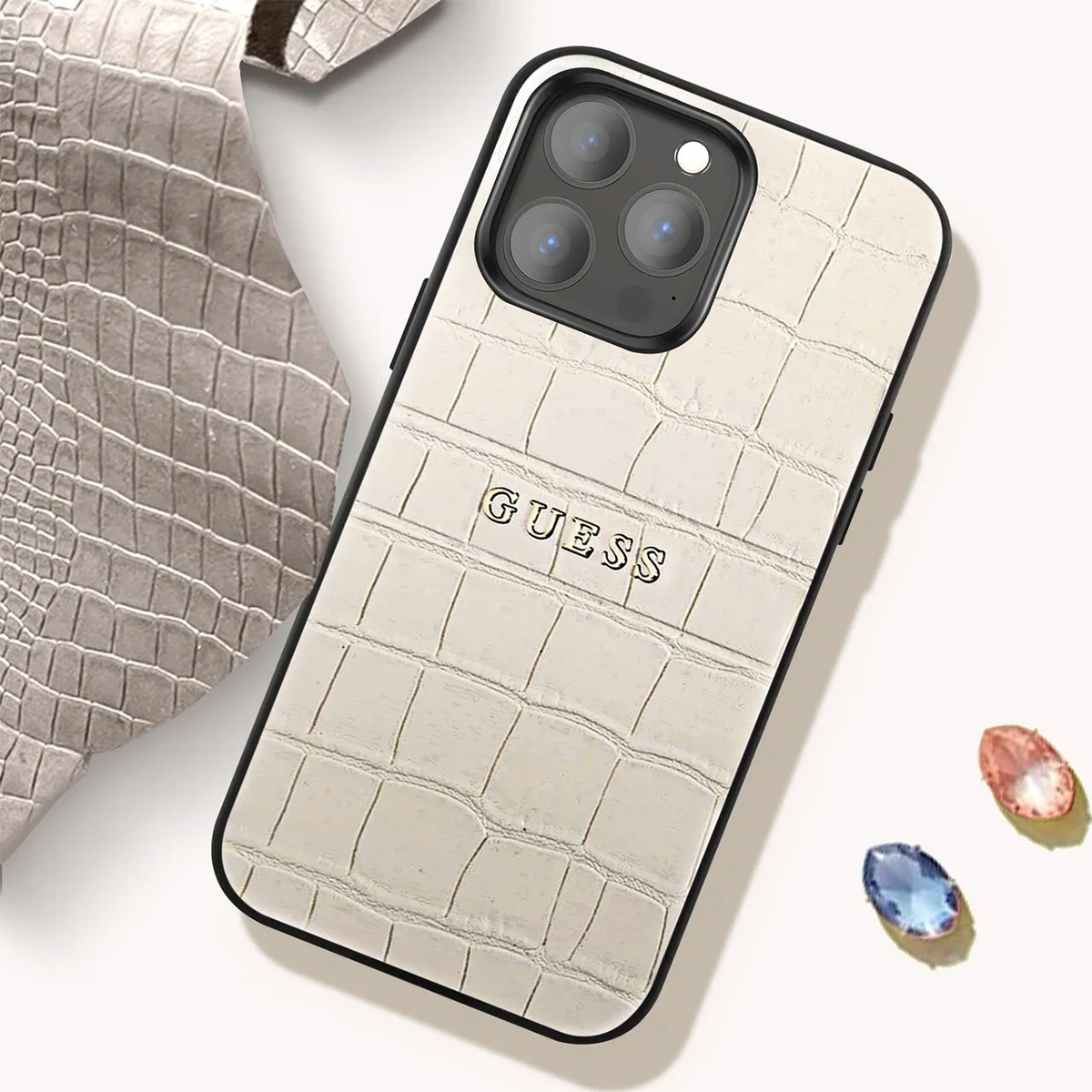 Una custodia per telefono Guess bianca con motivo coccodrillo. La custodia ha una cornice nera. Sono visibili due gemme. Un pezzo di pelle grigia è sul lato.
