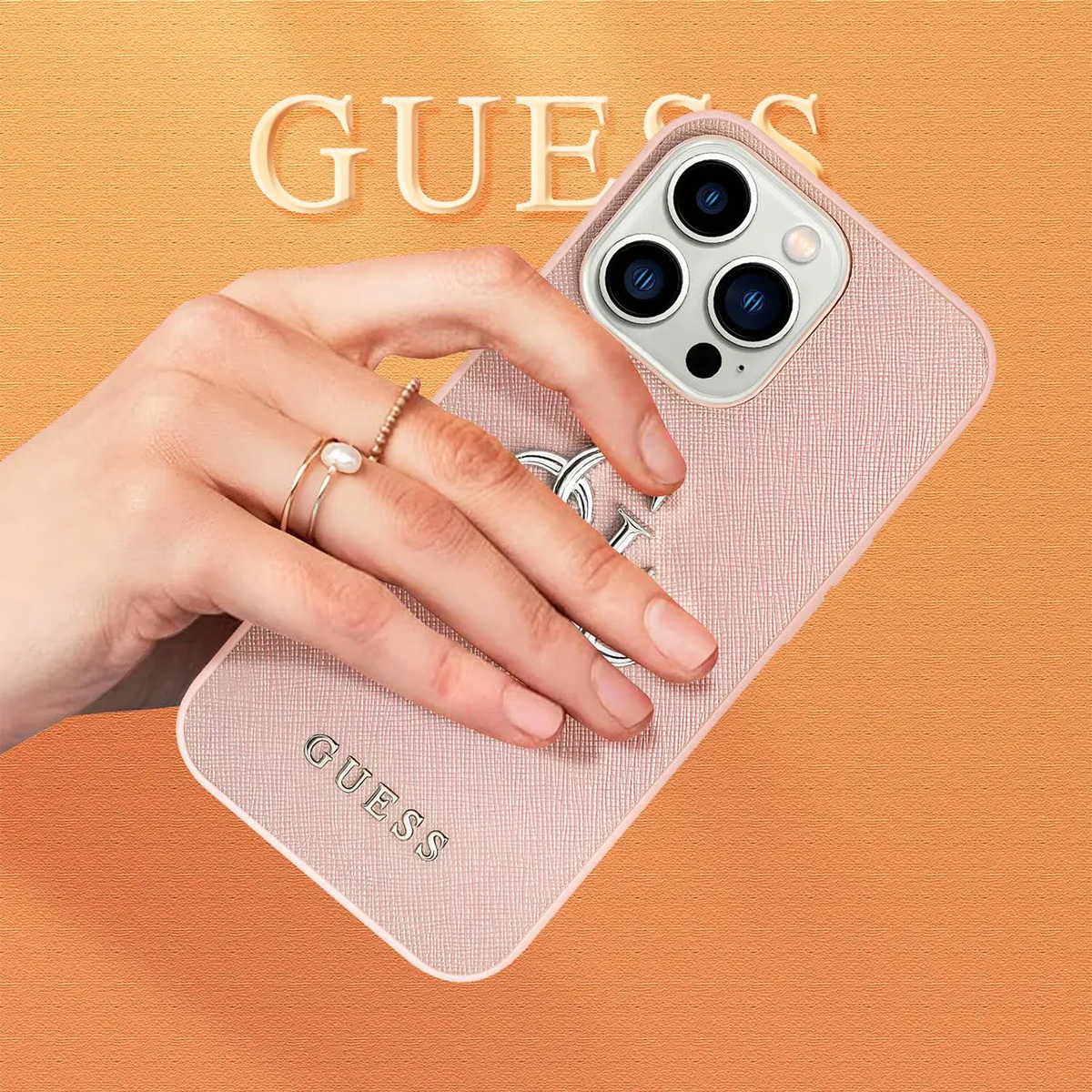 Una mano che tiene una custodia per telefono Guess rosa con un anello d'argento. Lo sfondo è arancione con il logo Guess.