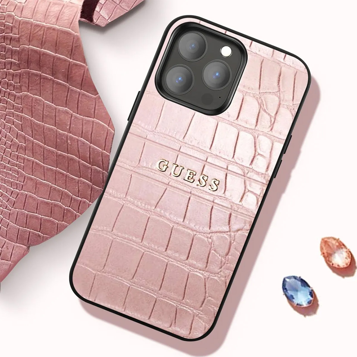 Custodia per telefono Guess rosa con texture coccodrillo. La custodia è su una superficie bianca, con due gemme e un pezzo di texture coccodrillo.