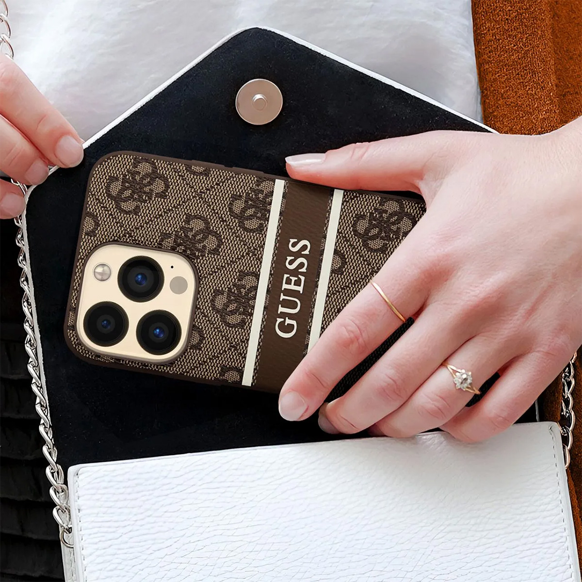Una persona tiene una custodia per telefono Guess in una borsa bianca. La custodia è marrone con un motivo logo e una striscia bianca con "GUESS".