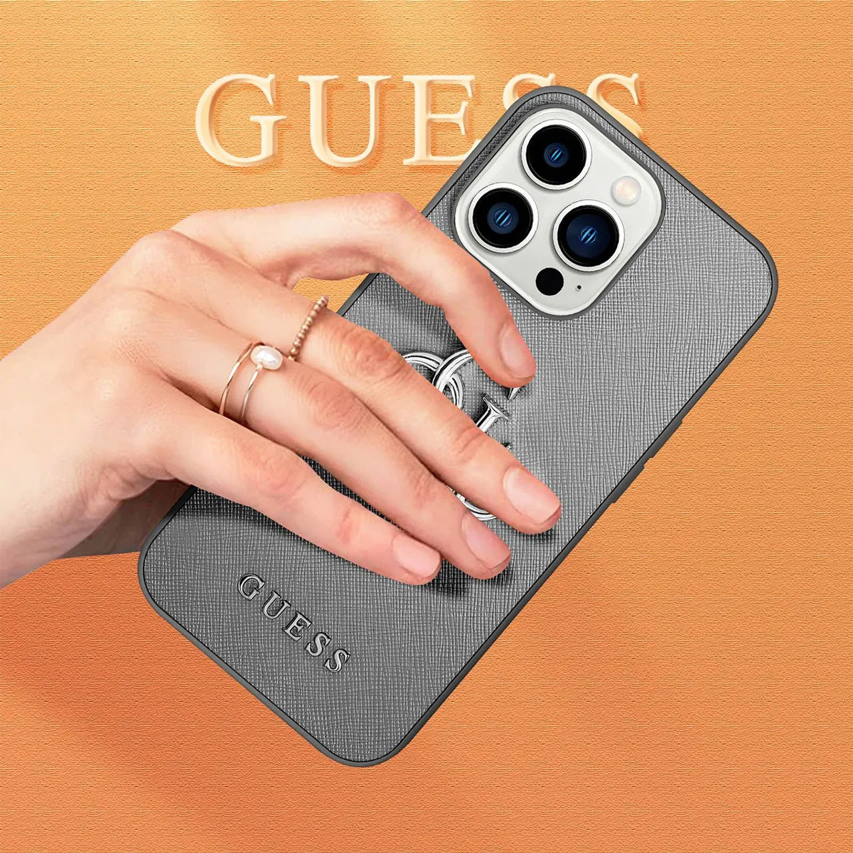 Una mano tiene una custodia grigia con il logo Guess. Lo sfondo è arancione con la scritta GUESS. Ci sono anelli sulle dita.