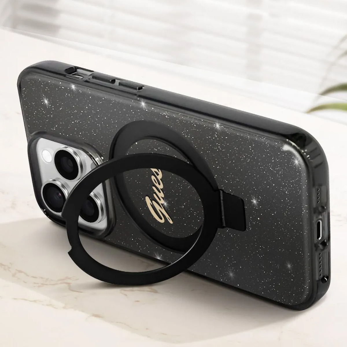 Una custodia nera per telefono con supporto ad anello. La custodia ha una finitura glitterata. Il supporto ha la scritta 'Guess'.