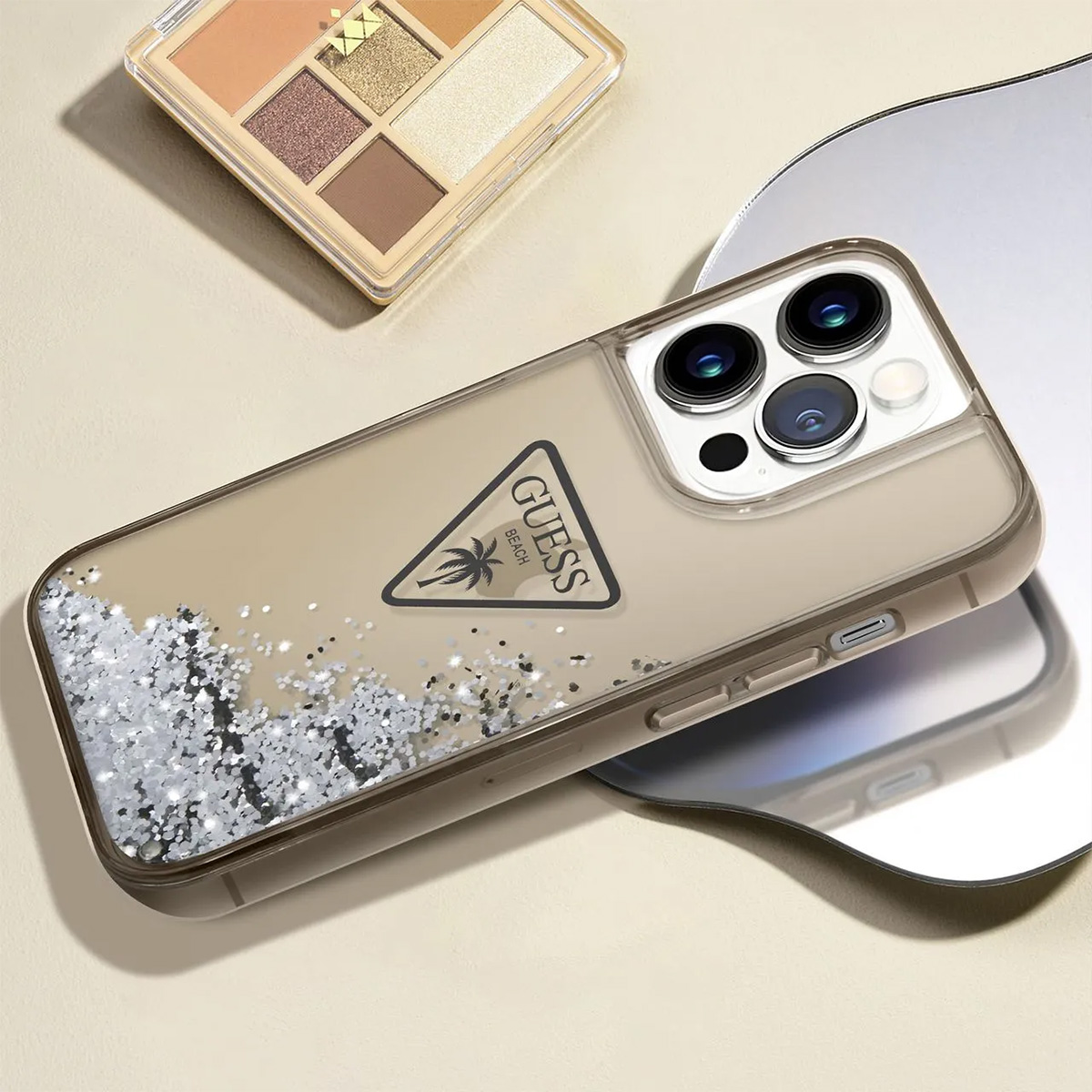 Una custodia per telefono con logo Guess, palette ombretti e specchio su superficie beige.