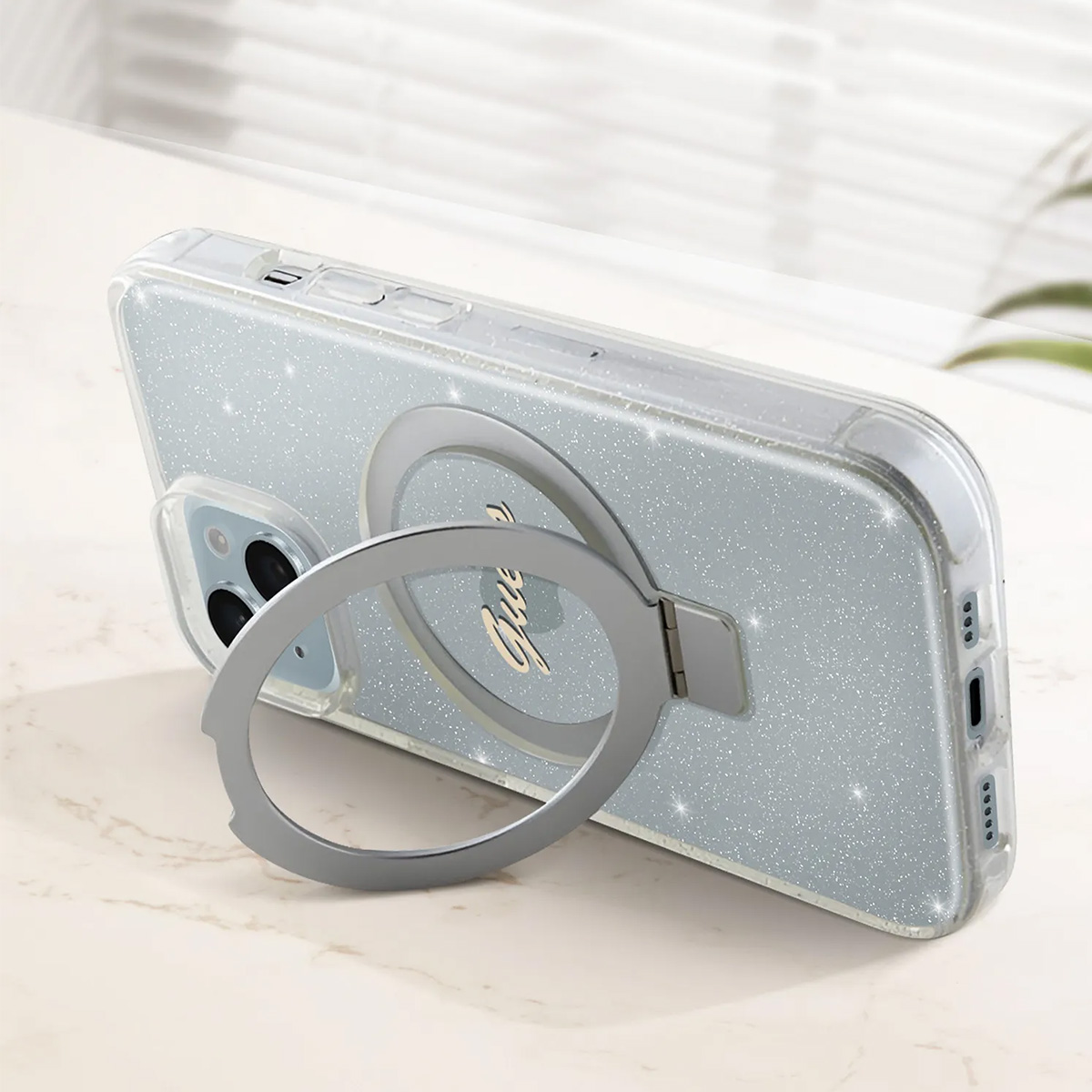 Un telefono in una custodia glitterata con un supporto ad anello argentato. L'anello è attaccato a un cerchio con la scritta 'Guess'.