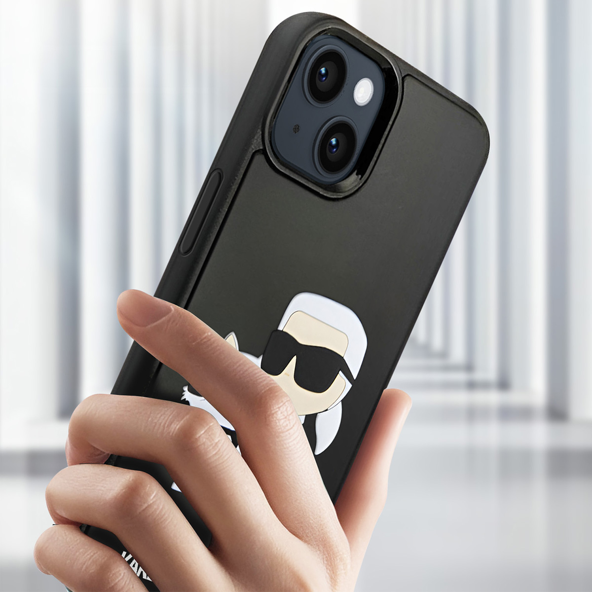 Una mano tiene in mano un telefono nero con una custodia Karl Lagerfeld. La custodia ha un'immagine a cartoni animati di Karl. Lo sfondo è sfocato.