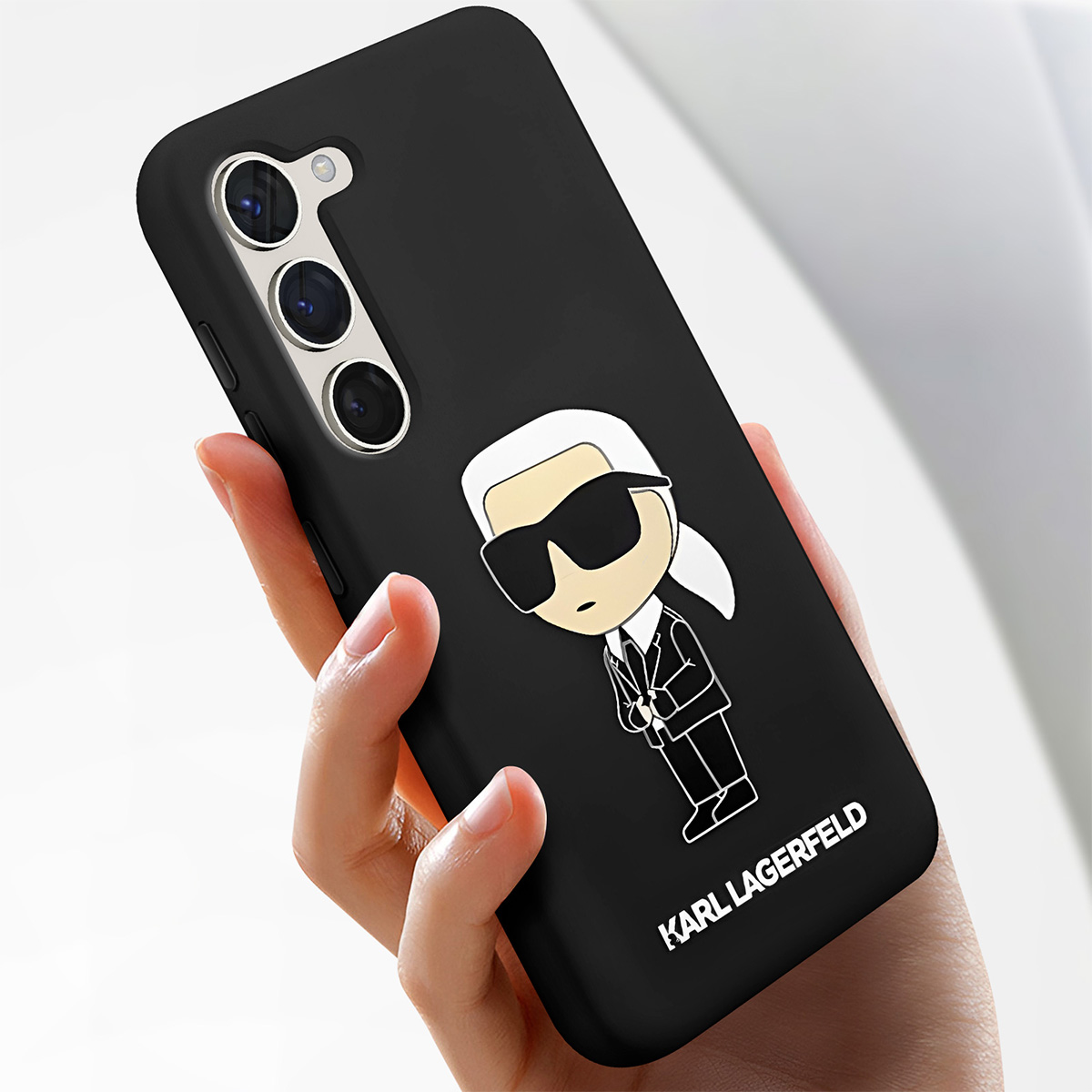 Una mano tiene una custodia nera per telefono. Ha un design a cartone animato di Karl Lagerfeld e il nome del marchio. Il telefono ha tre fotocamere.