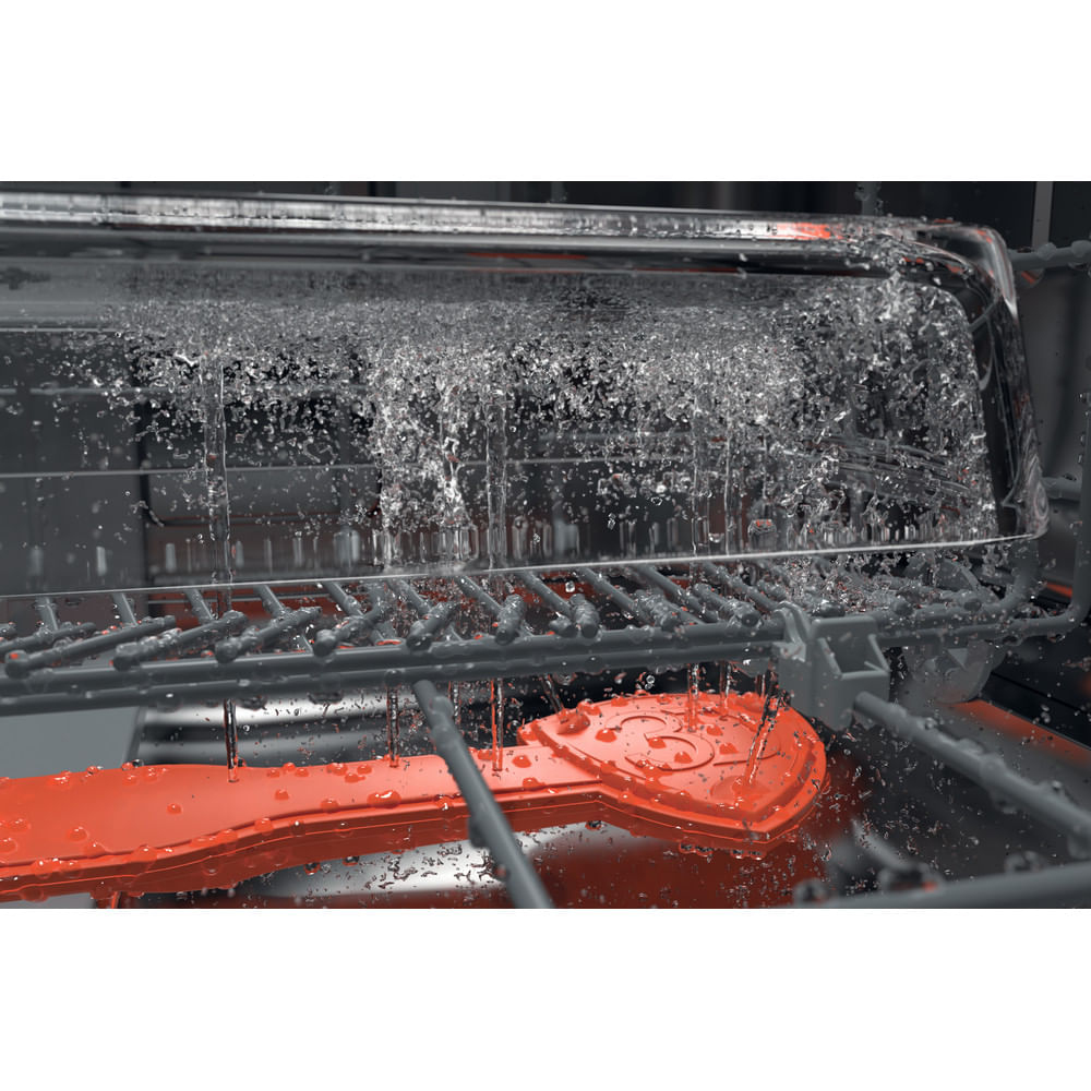 Dentro de un lavavajillas, el agua se pulveriza hacia abajo. Un objeto rojo en forma de corazón está en la parte inferior. Se ven rejillas grises y un recipiente transparente.