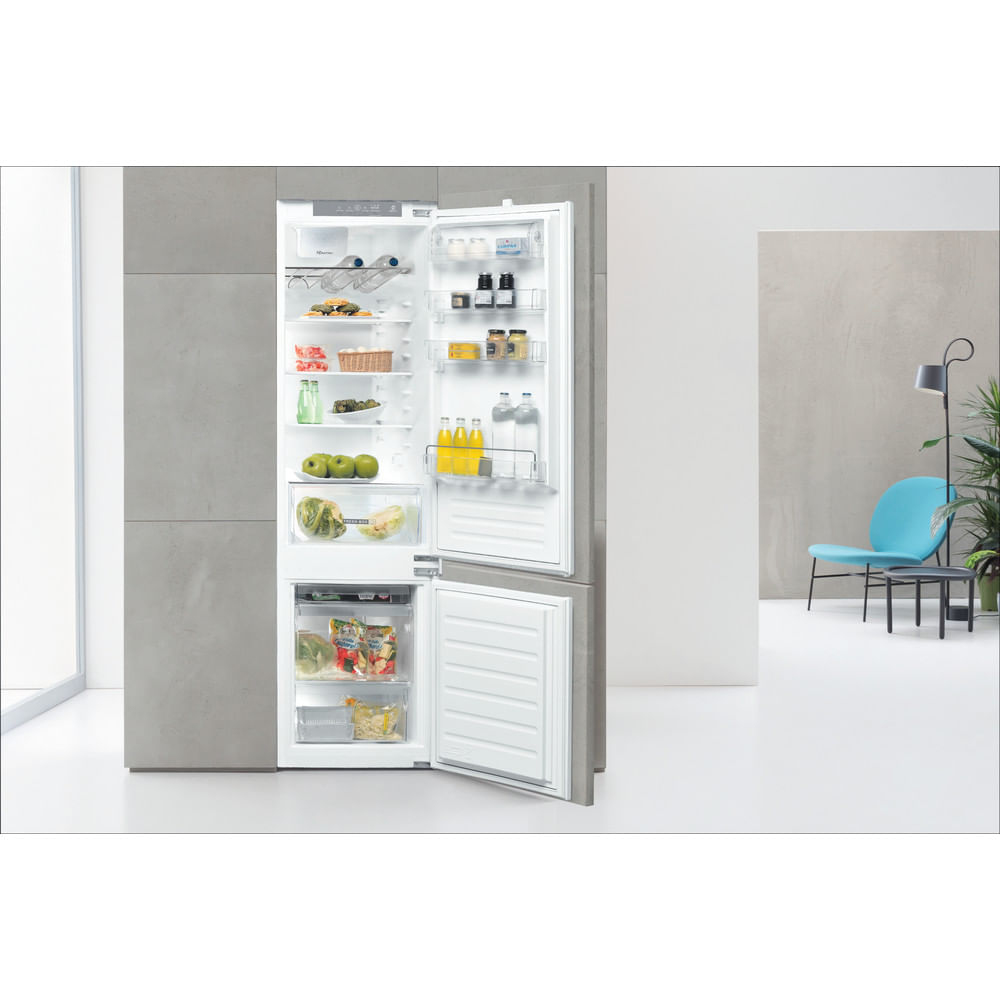 Open fridge with food: apples, vegetables, drinks. Blue chair and lamp on the right. Gray wall on the left.