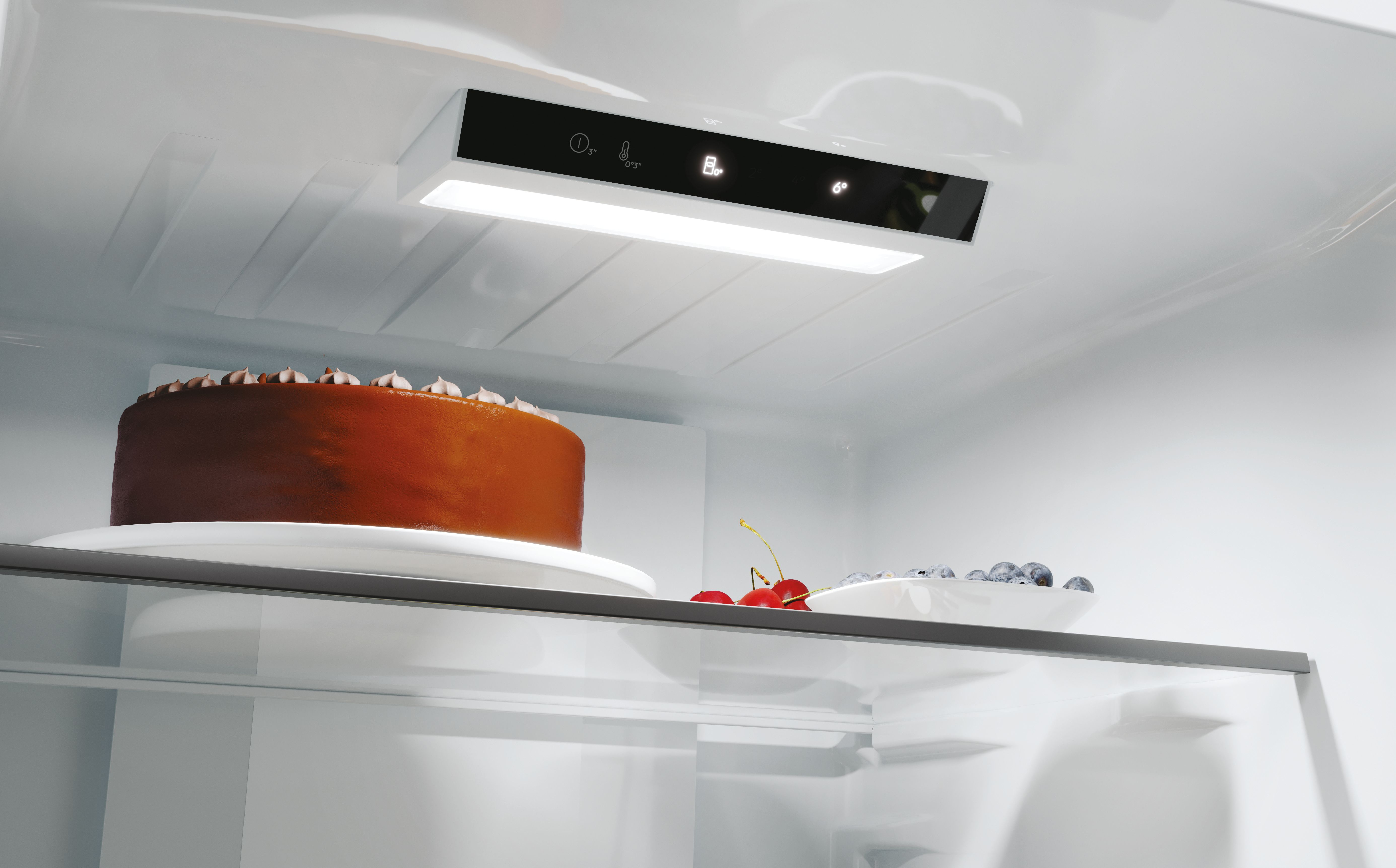 Dentro de un refrigerador, un pastel de chocolate en un plato, cerezas y arándanos en un cuenco y un panel de control.