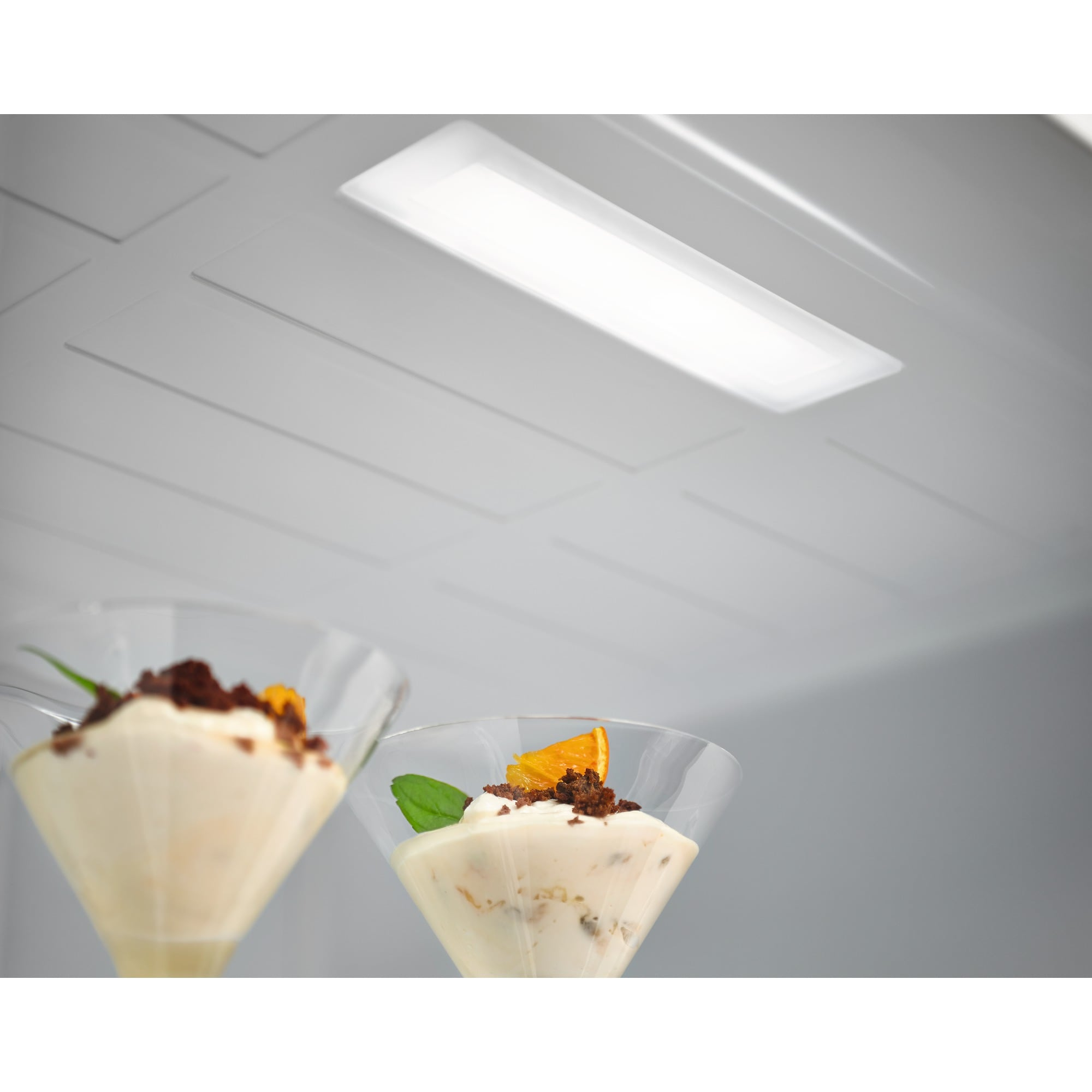 Dos postres en vasos transparentes bajo una luz brillante. Cada postre tiene una guarnición y una rodaja de naranja.