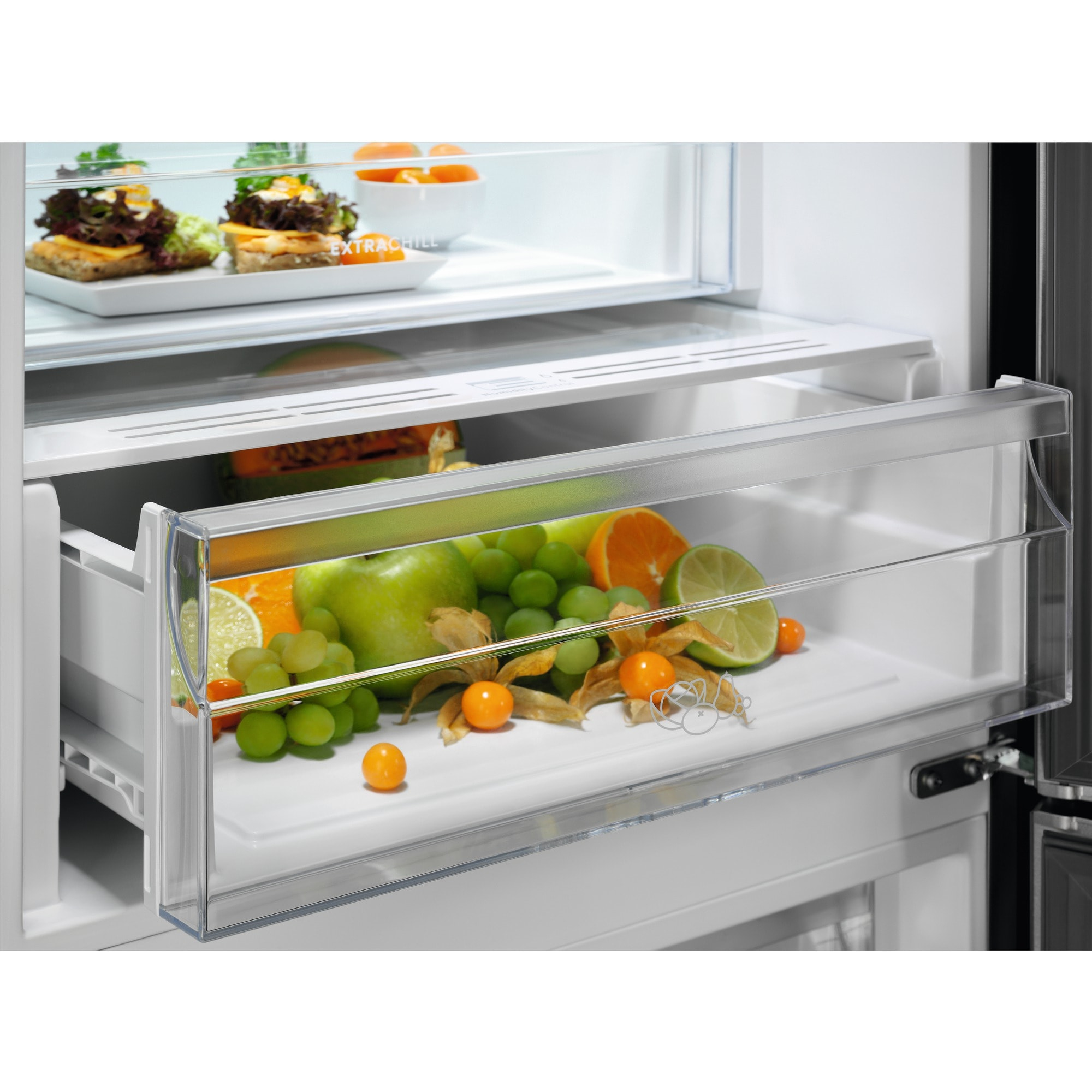 Un cajón de nevera con frutas como uvas verdes, manzanas, naranjas y physalis. Sándwiches en el estante superior.