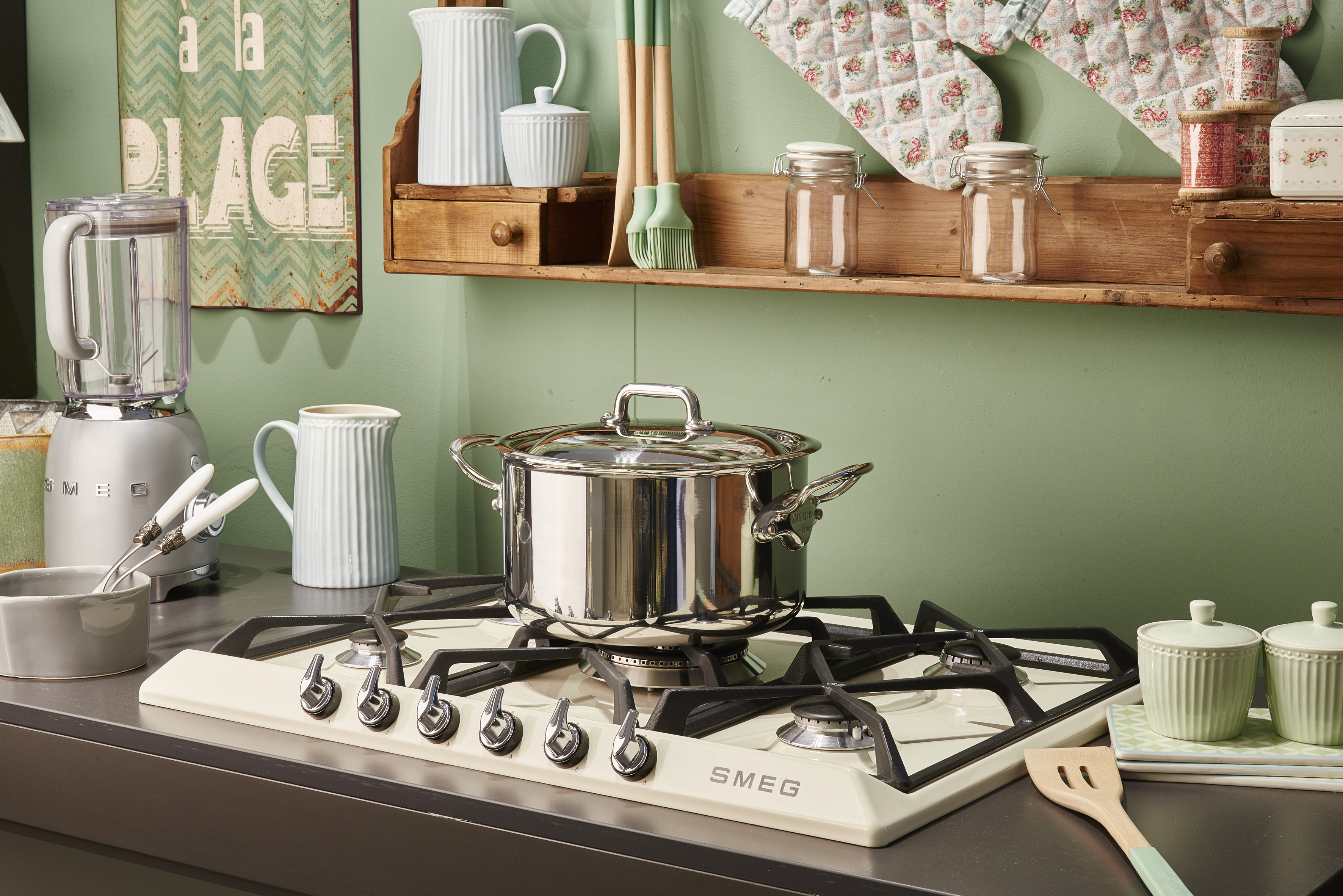 Una escena de cocina con una estufa, una olla y frascos en un estante. A la izquierda hay una licuadora y un letrero en la pared.