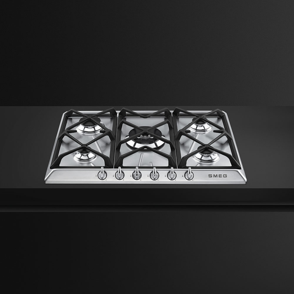 Una placa de gas Smeg de acero inoxidable con cinco quemadores. Soportes y mandos negros, sobre fondo oscuro.
