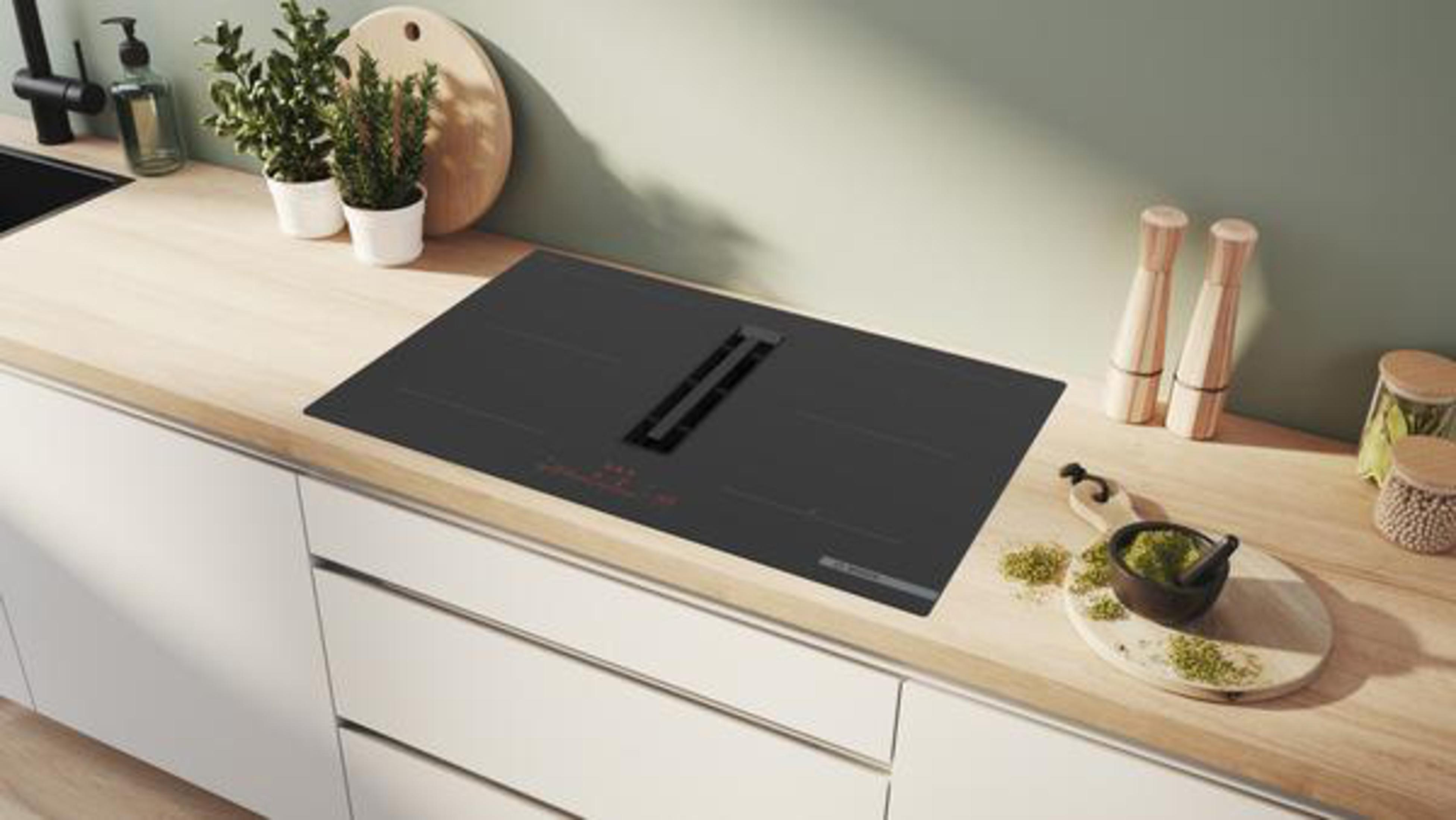 Un comptoir de cuisine moderne avec une table de cuisson noire, des plantes et des ustensiles de cuisine. Meubles blancs en dessous et mur vert clair.
