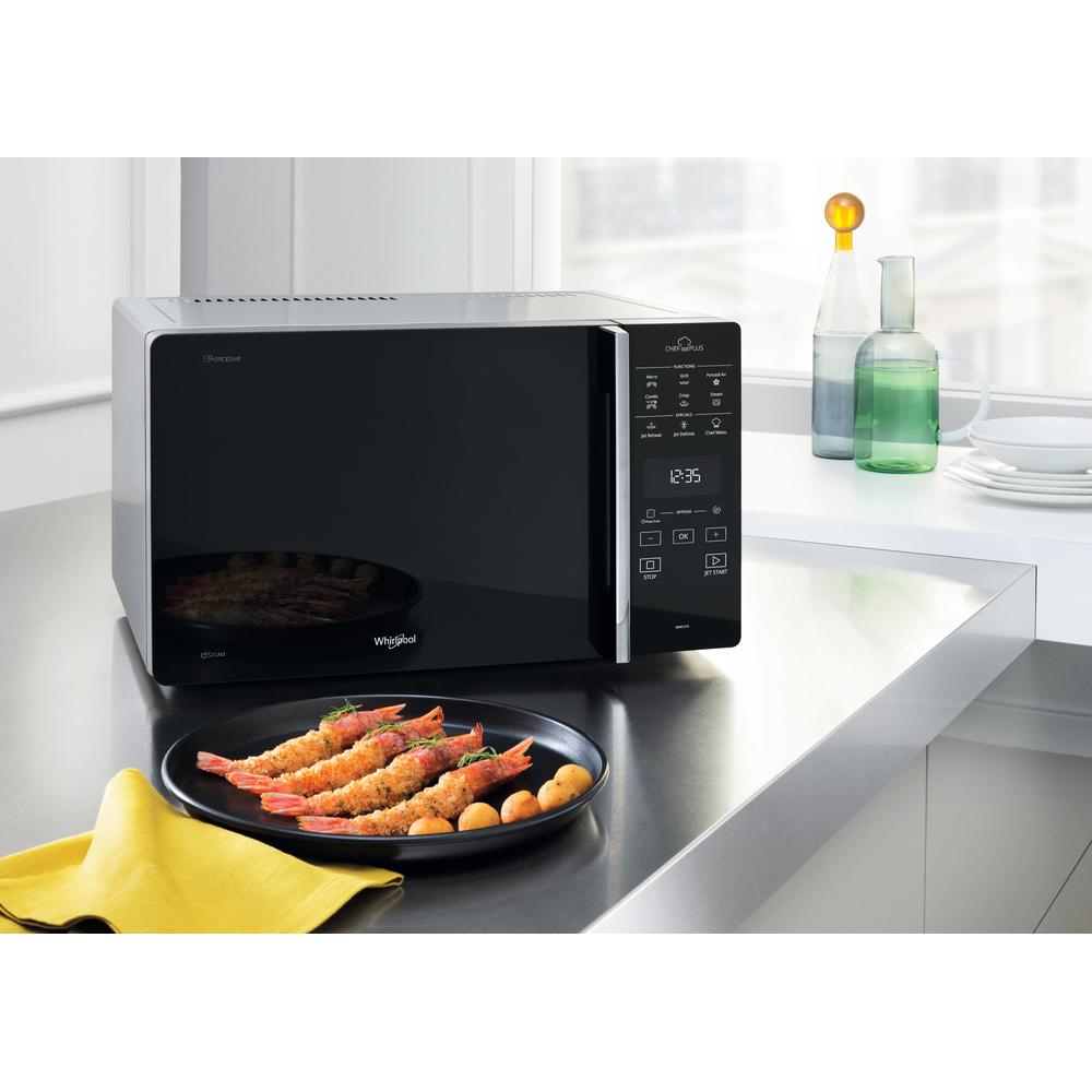 Un forno a microonde nero su un bancone. Un piatto di gamberi e patate in primo piano. Bottiglie sullo sfondo.