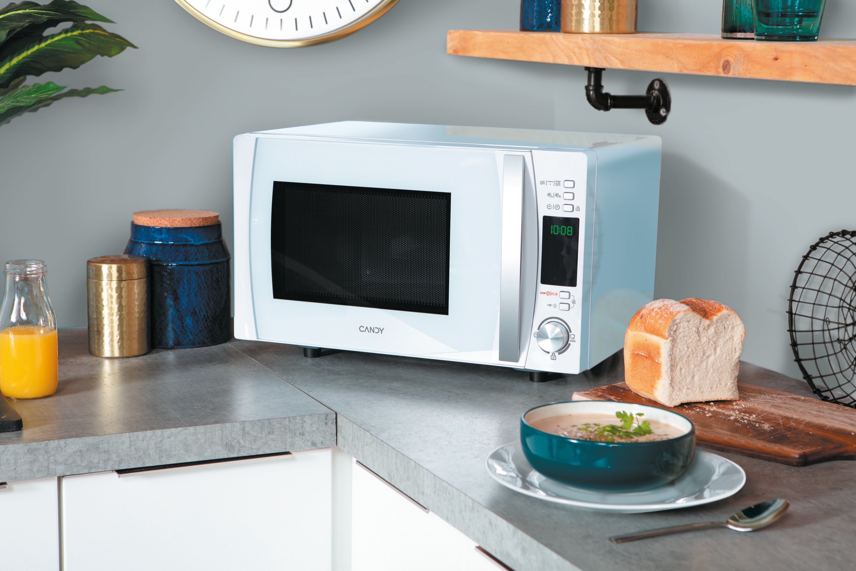 Un horno microondas blanco sobre una encimera, con pan, sopa y frascos. Hay un reloj en la pared.