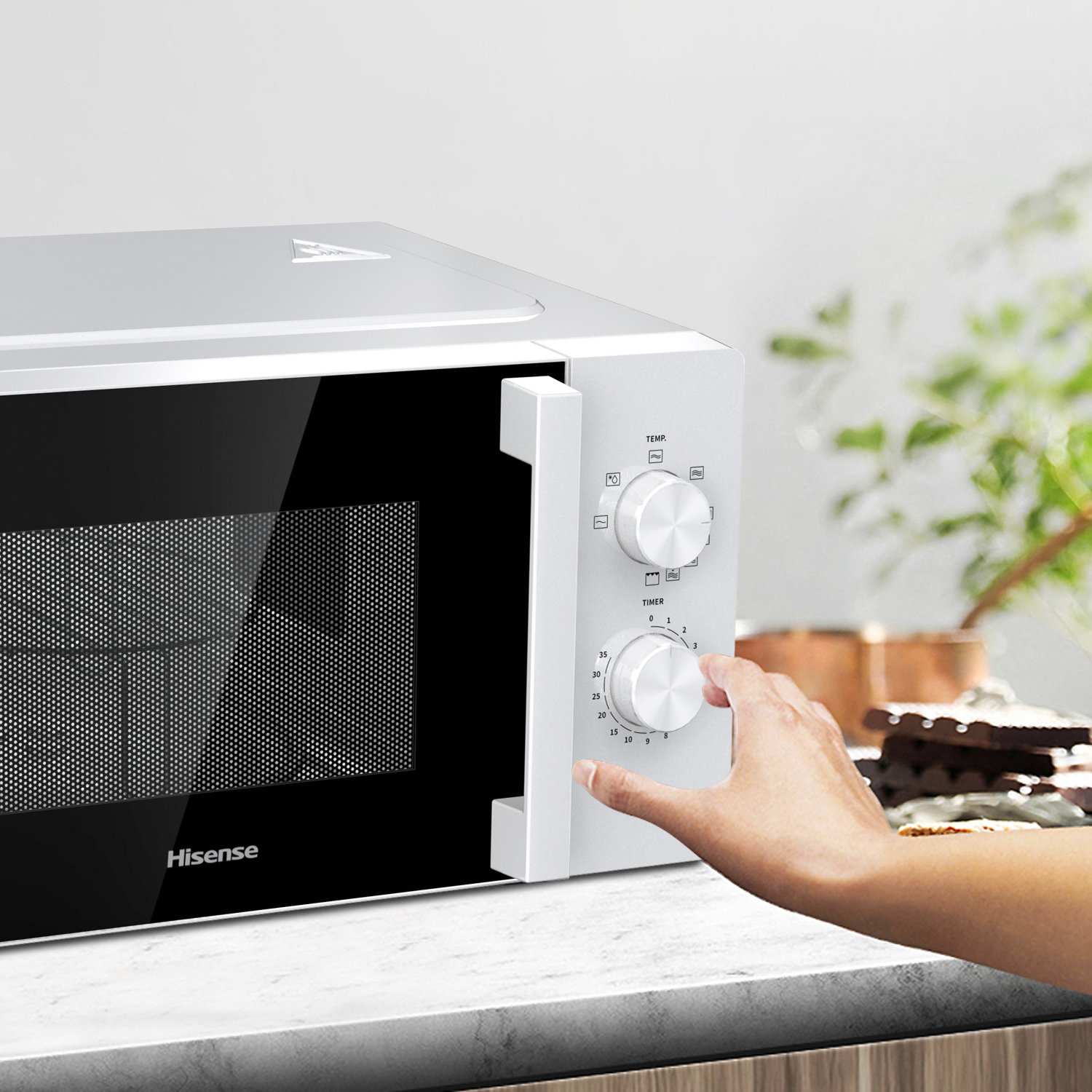 Ein weißer Hisense-Mikrowellenherd, bei dem eine Hand den Timer-Knopf dreht. Der Hintergrund zeigt verschwommenes Grün und Lebensmittel.